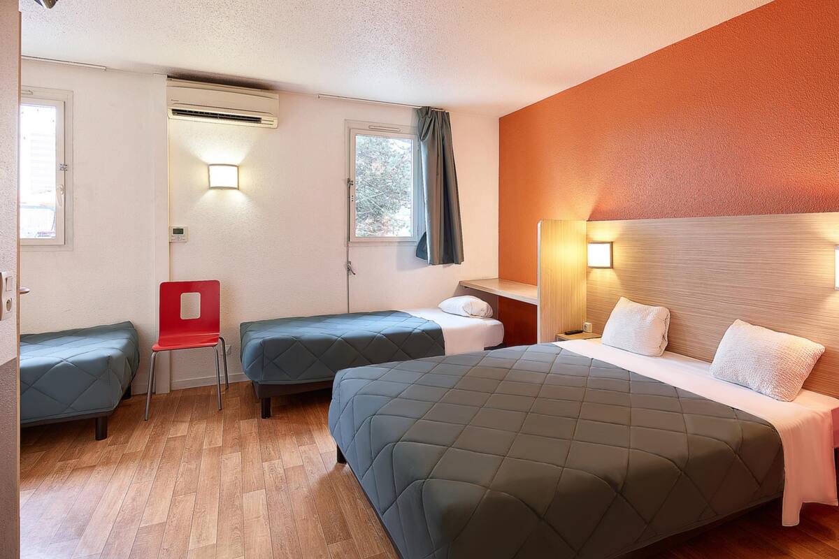 A hotel room with two beds, a red chair, a window with curtains, and an air conditioning unit.