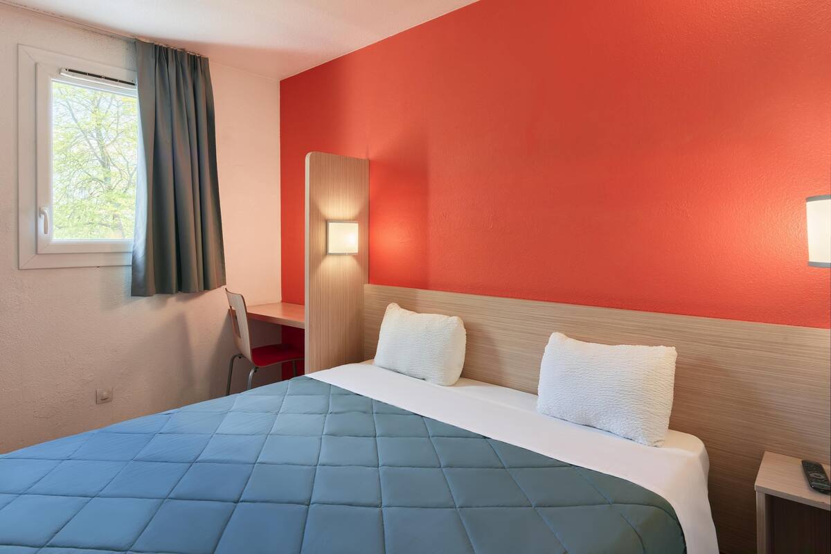 A hotel room with a bed, a desk, a chair, a nightstand, and a window with curtains.