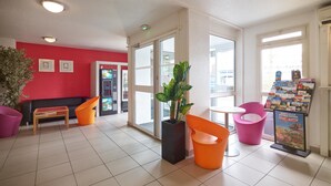 A hotel lobby with a red wall, a vending machine, a seating area with orange chairs, a potted plant, and a poster.