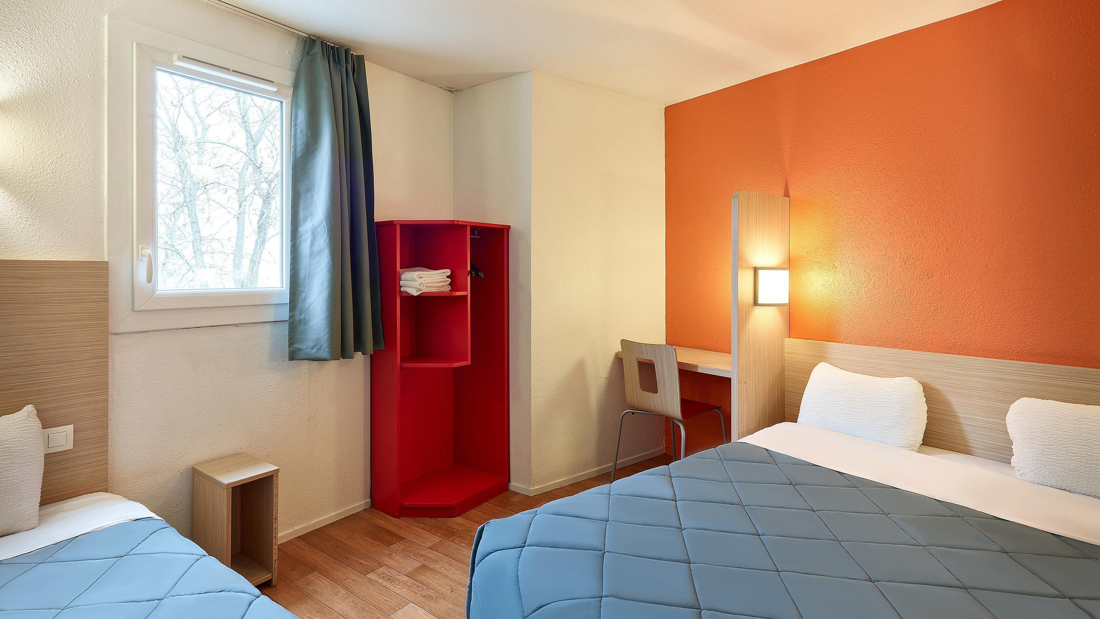 Une chambre d’hôtel avec deux lits, un bureau, une armoire rouge et une fenêtre avec des rideaux.