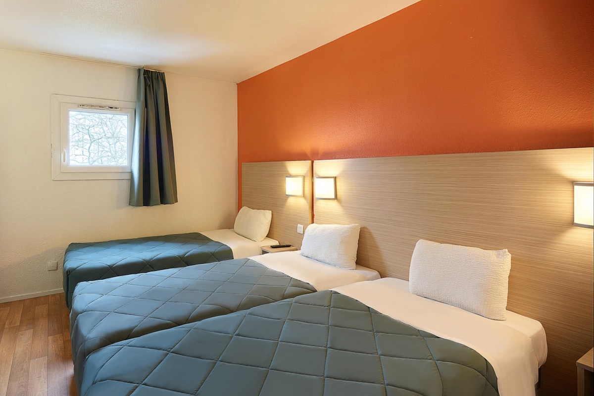 A hotel room with two beds, a window with curtains, a wall-mounted light, and a wooden floor.