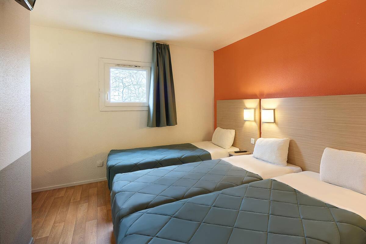 A hotel room with two beds, a window with curtains, a wall-mounted light, and a wooden floor.