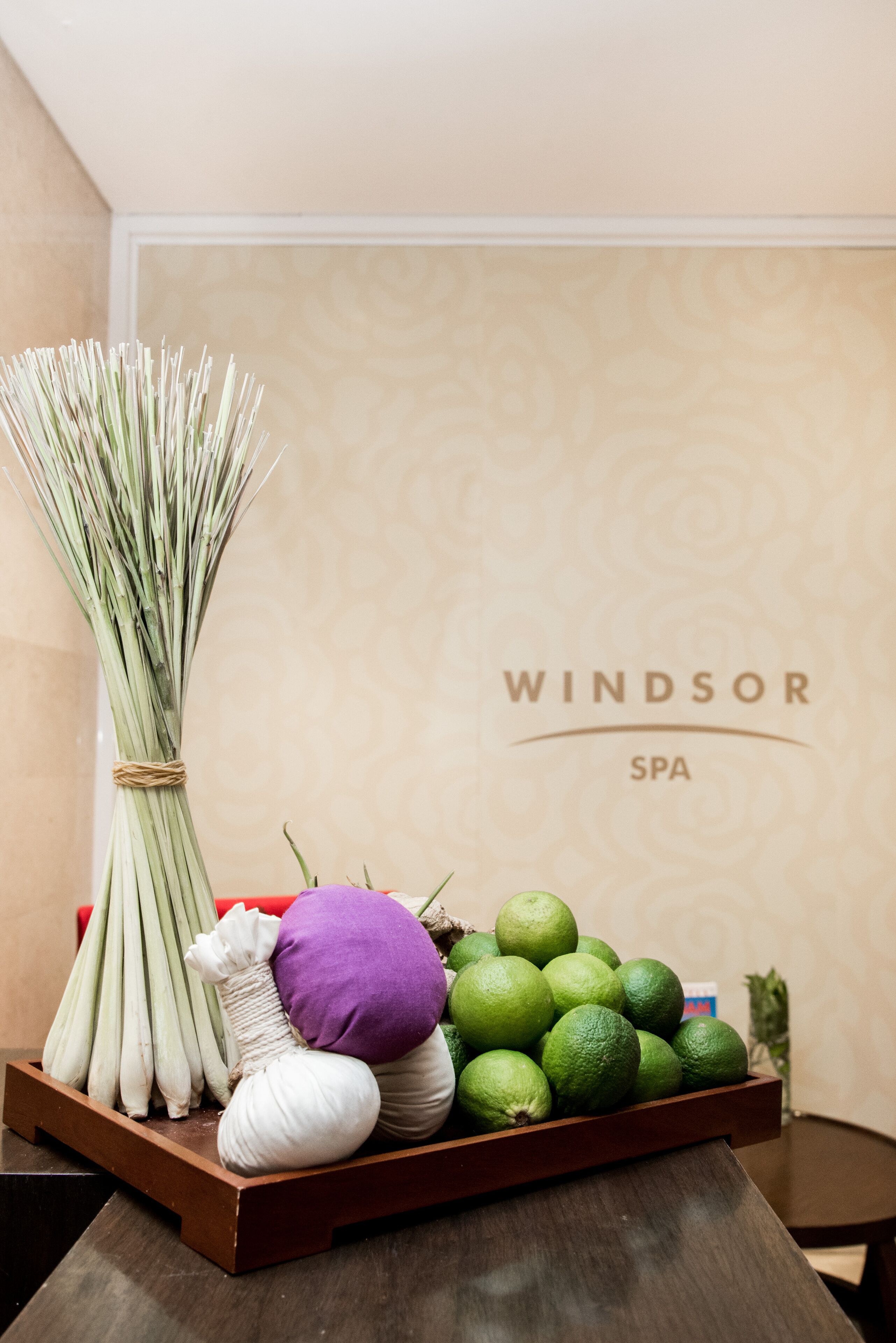 A wooden tray with fresh herbs, a purple pillow, and green limes on a table in a room with a Windsor Spa sign in the background.