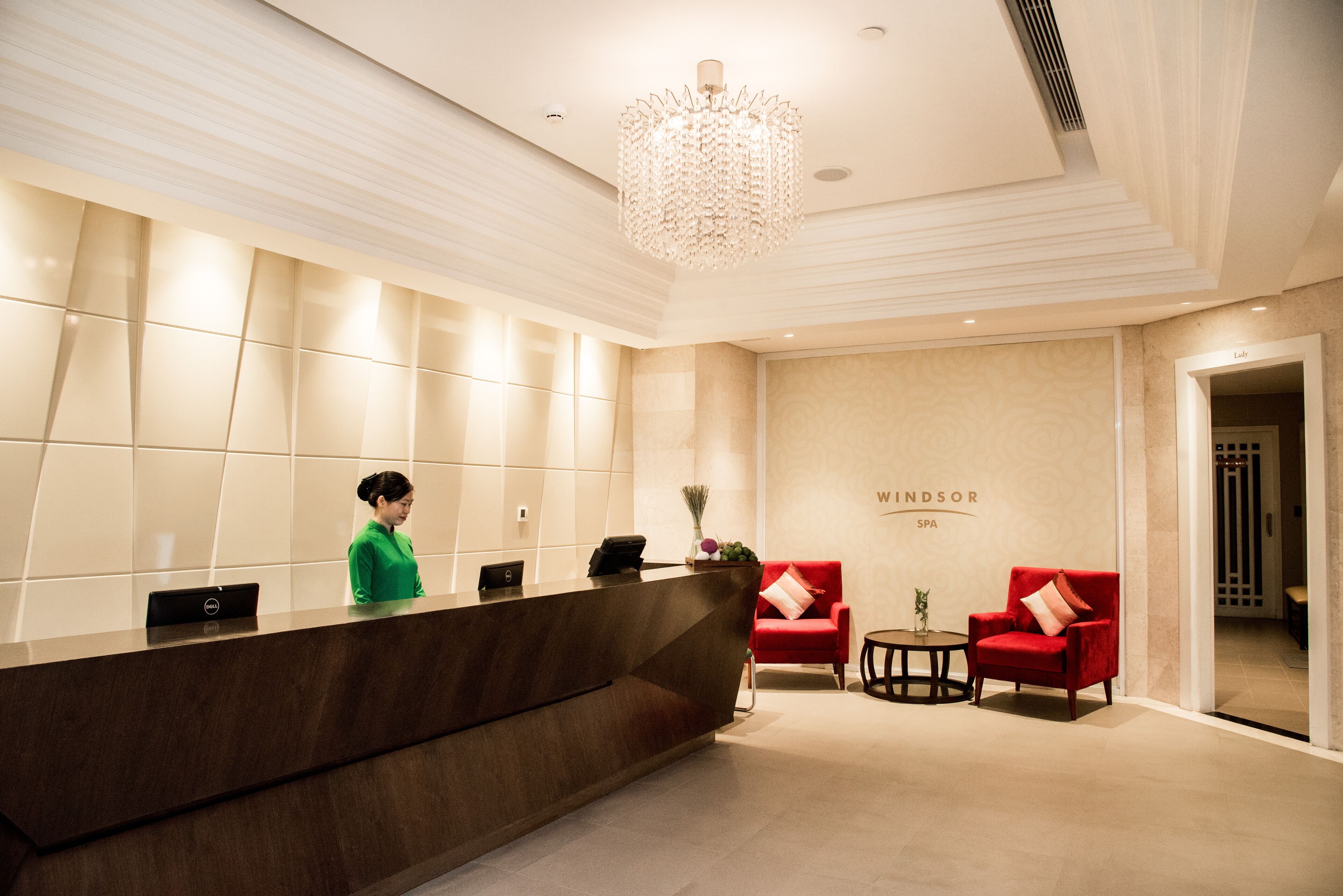 A reception area with a wooden reception desk, a receptionist, two red armchairs, a small table with a vase, and a chandelier.