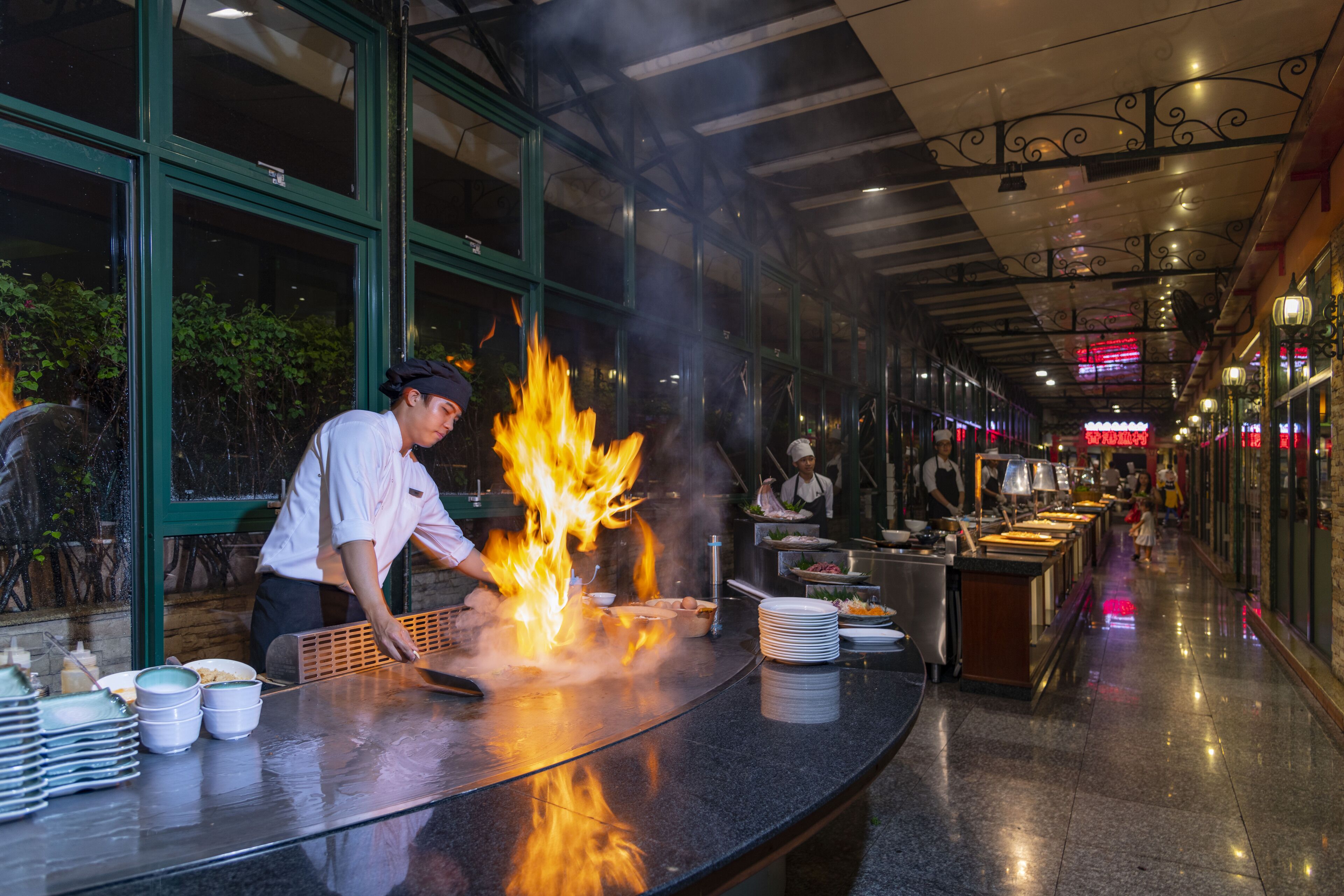 Un chef realizando una técnica de cocción con fuego en la barra de un restaurante con una decoración moderna y vistas a la calle.