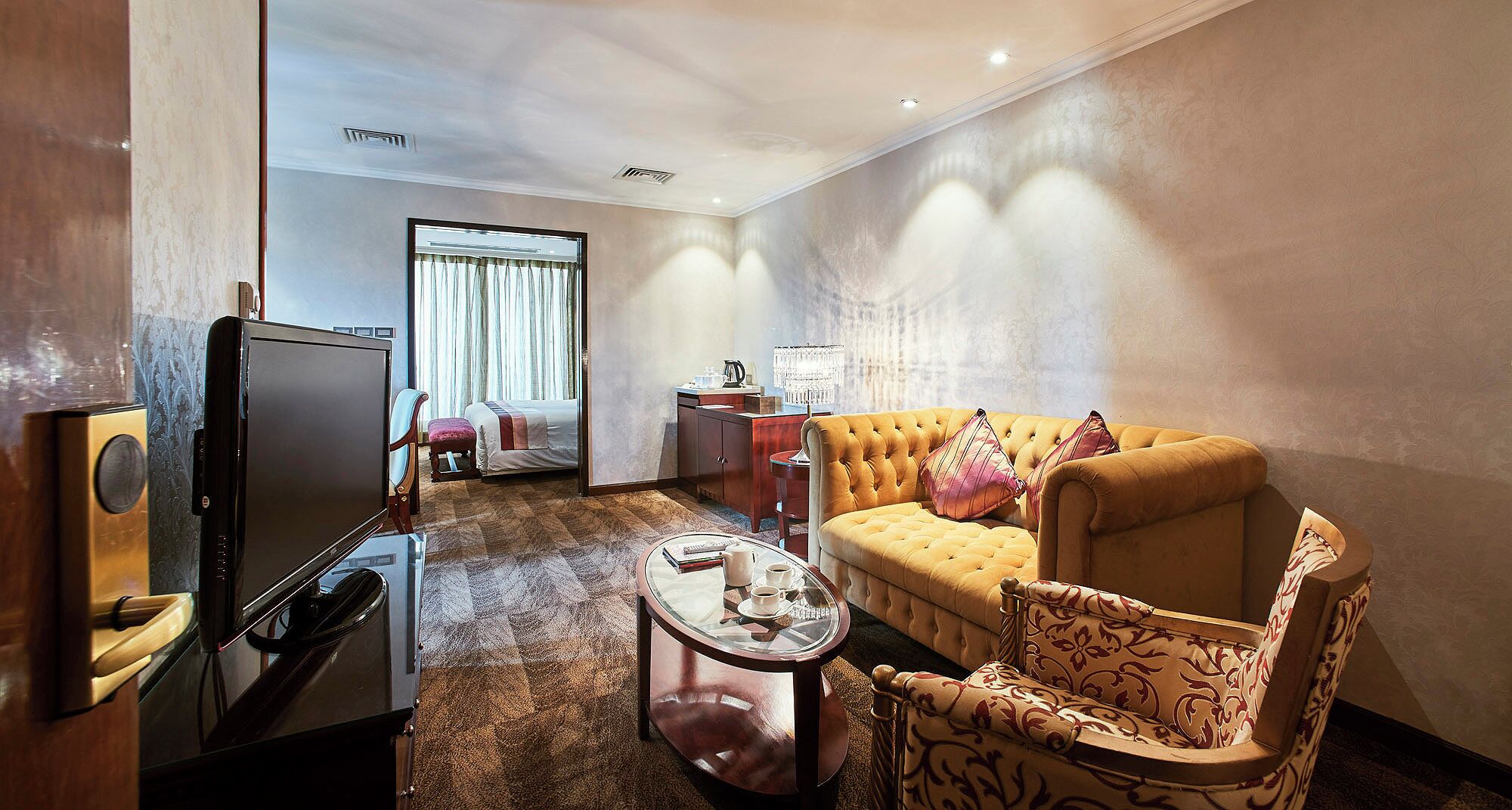 A hotel room with a television, a sofa, a coffee table, a chair, a bedside table, and a window with curtains.