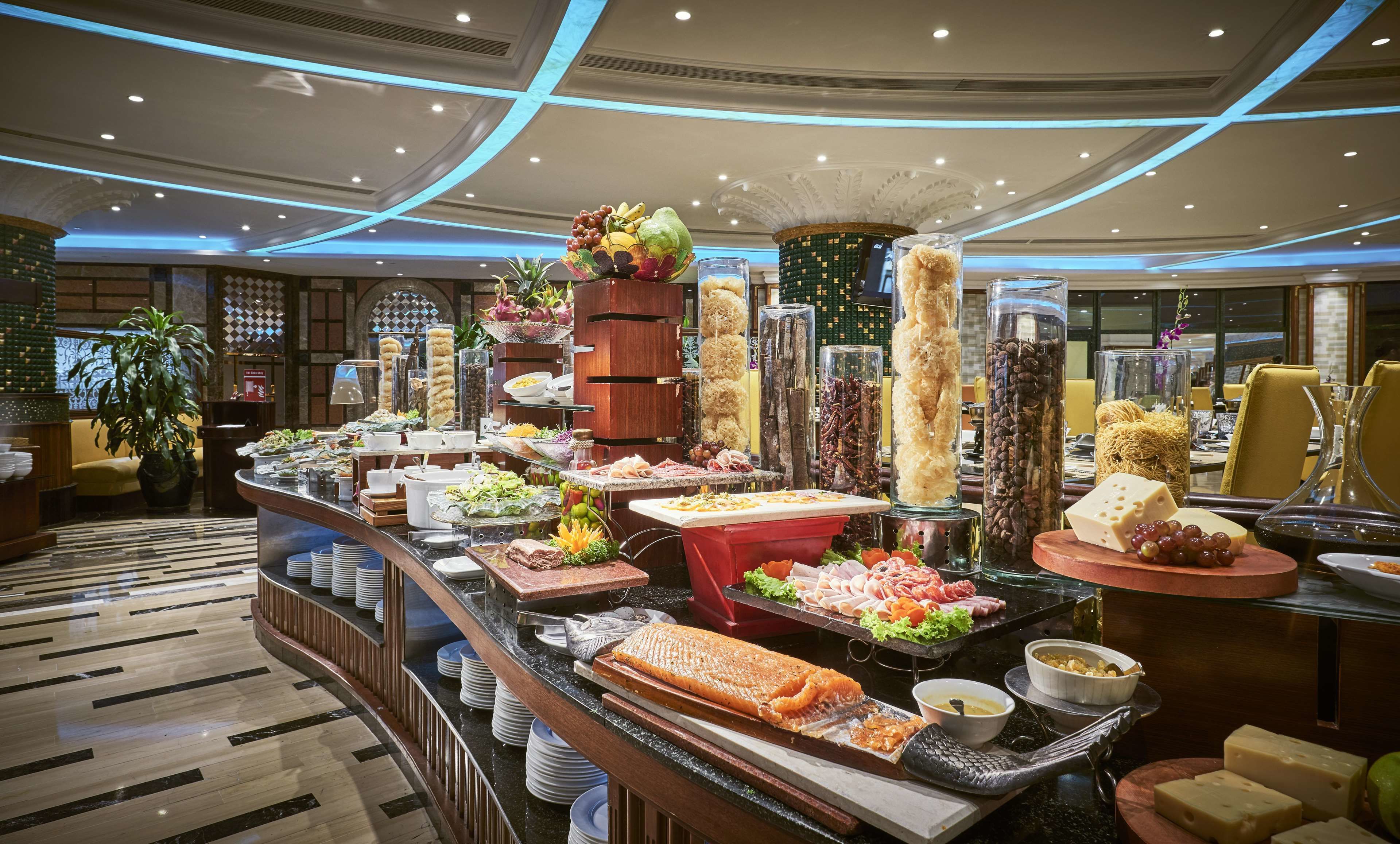 A buffet with a variety of food items, including meats, cheeses, and fruits, in a well-lit room with a marble floor and decorative ceiling lights.