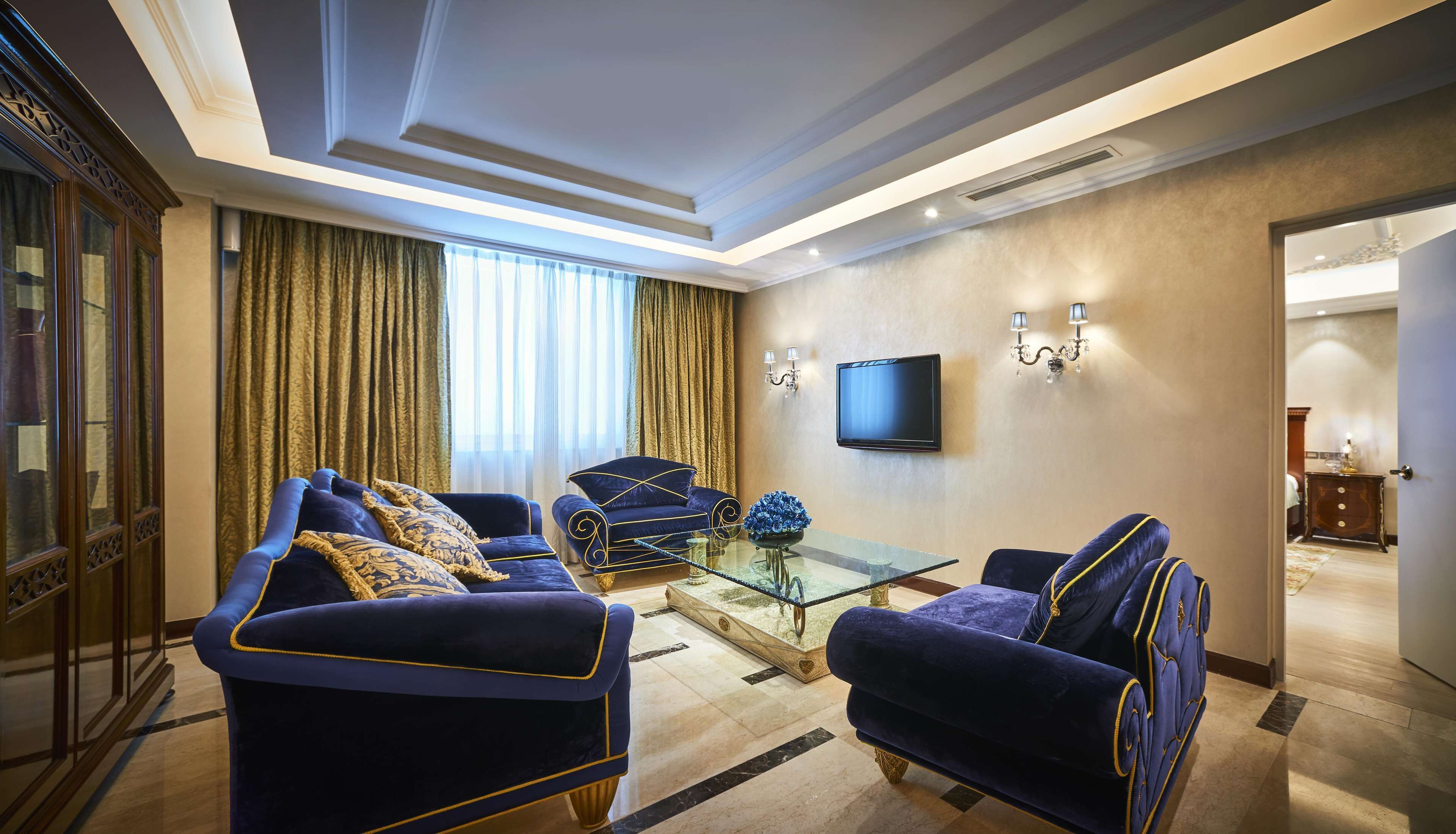 Una sala de estar lujosa con diseño moderno, que presenta un televisor de pantalla plana grande, elegantes sofás azules con adornos dorados y una mesa de centro de vidrio.