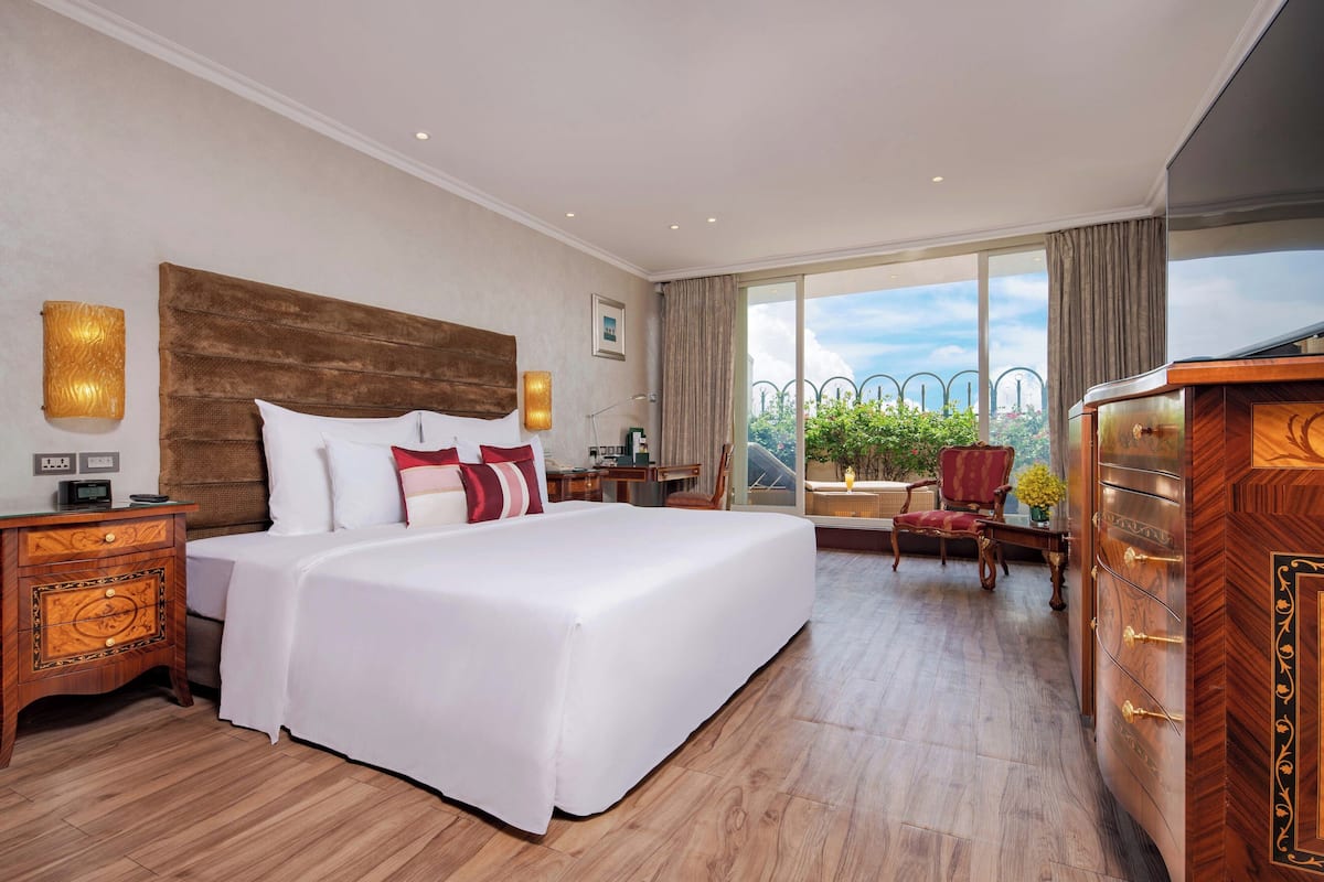 Une chambre d’hôtel avec un grand lit, des tables de chevet en bois, une commode et une vue sur la verdure grâce à de grandes fenêtres.