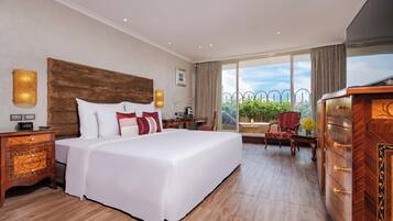 A hotel room with a large bed, wooden nightstands, a dresser, and a view of greenery through large windows.