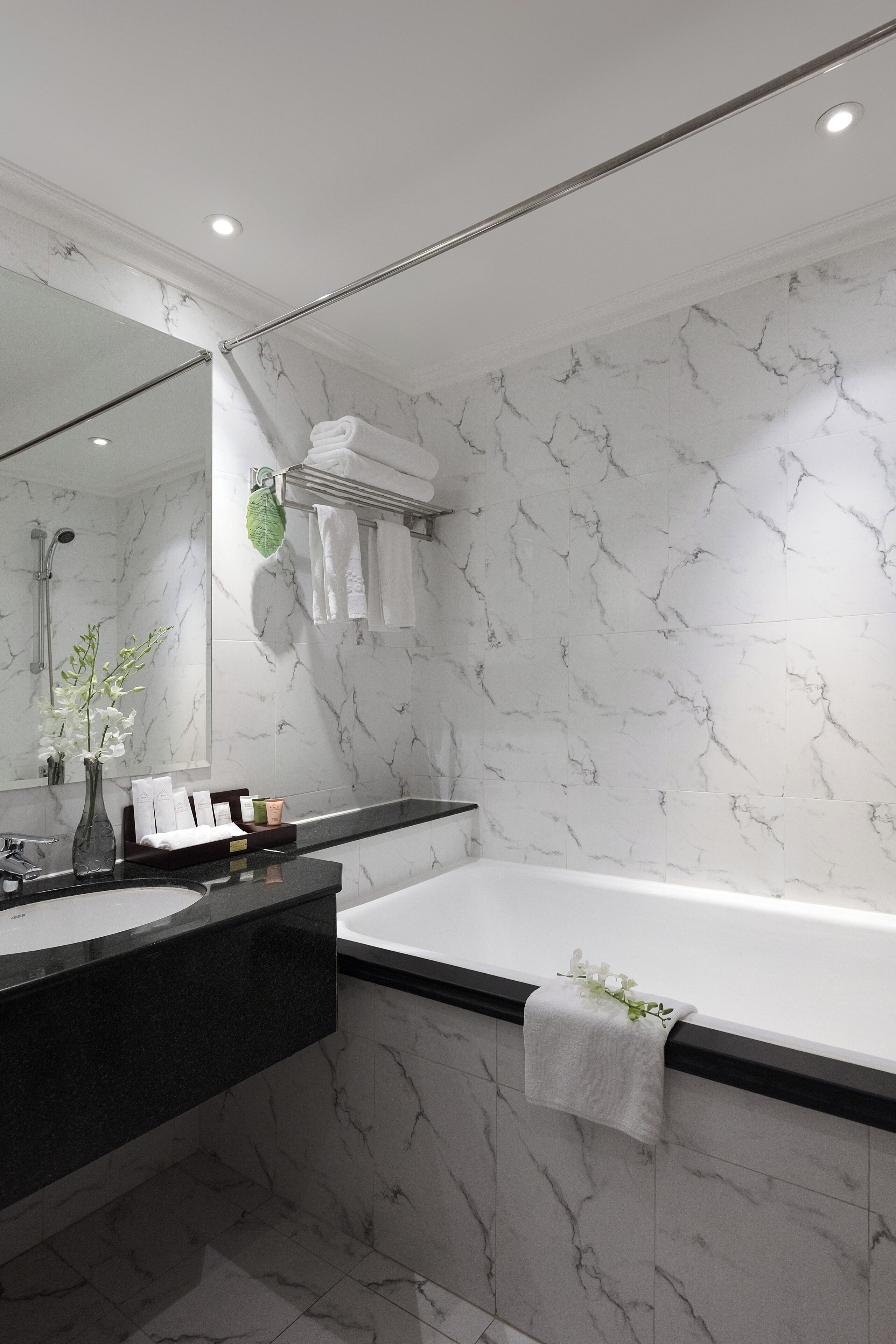 A modern bathroom with a marble backsplash, a large mirror, a black countertop, a white bathtub, and a towel rack with towels.