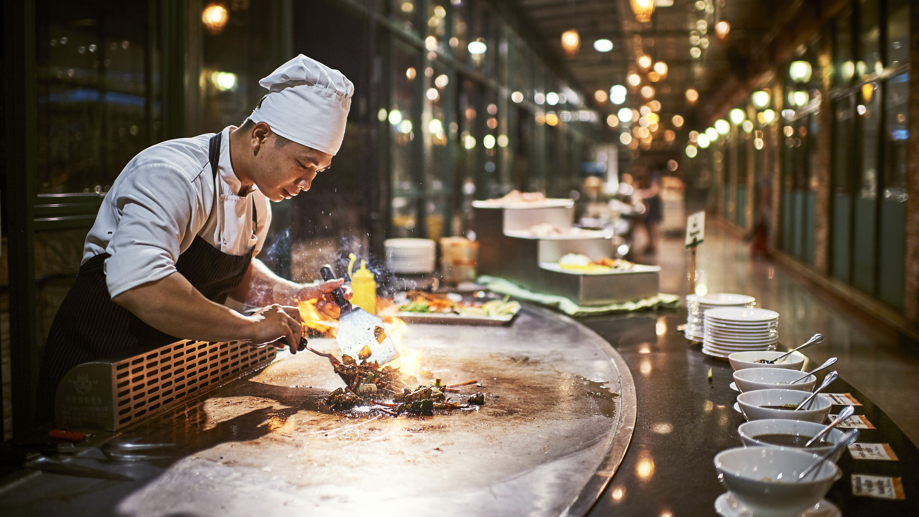 A chef cooking on a large, flat griddle in a well-lit kitchen with a long counter and multiple bowls of sauces lined up.