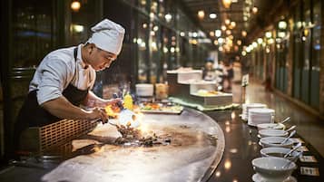 A chef cooking on a large, flat griddle in a well-lit kitchen with a long counter and multiple bowls of sauces lined up.