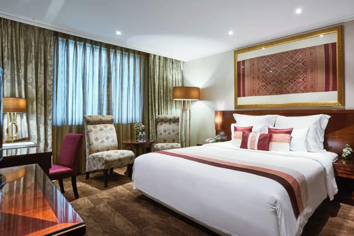 Une chambre d’hôtel avec un grand lit, deux fauteuils, un bureau avec une télévision et une grande fenêtre avec des rideaux.