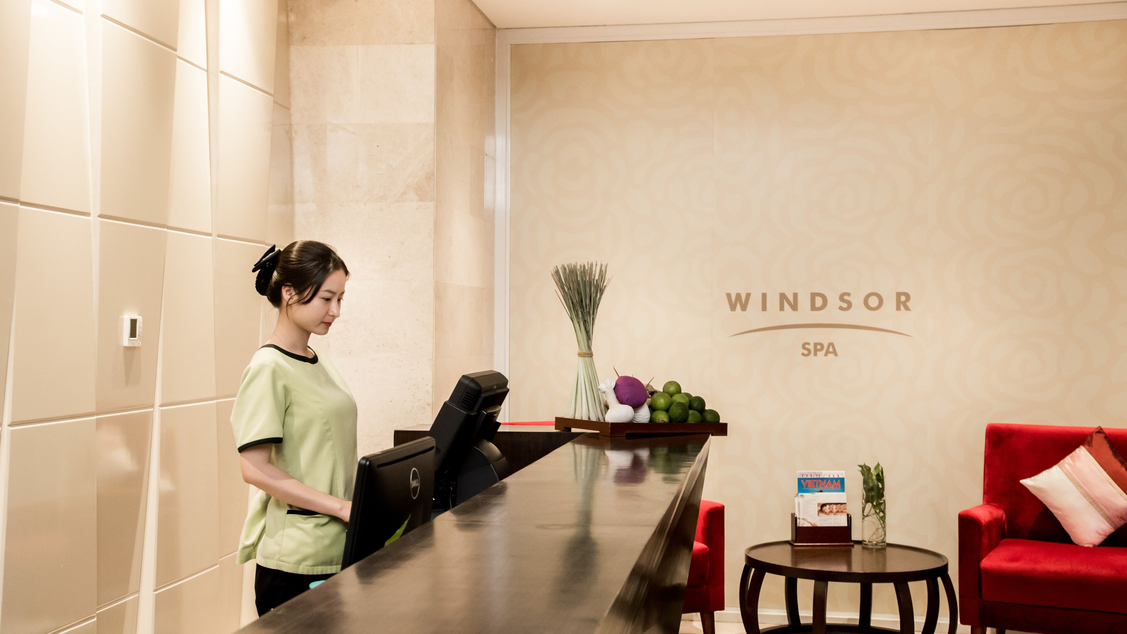 A reception desk with a computer, a vase with green plants, and a red sofa in a spa setting.