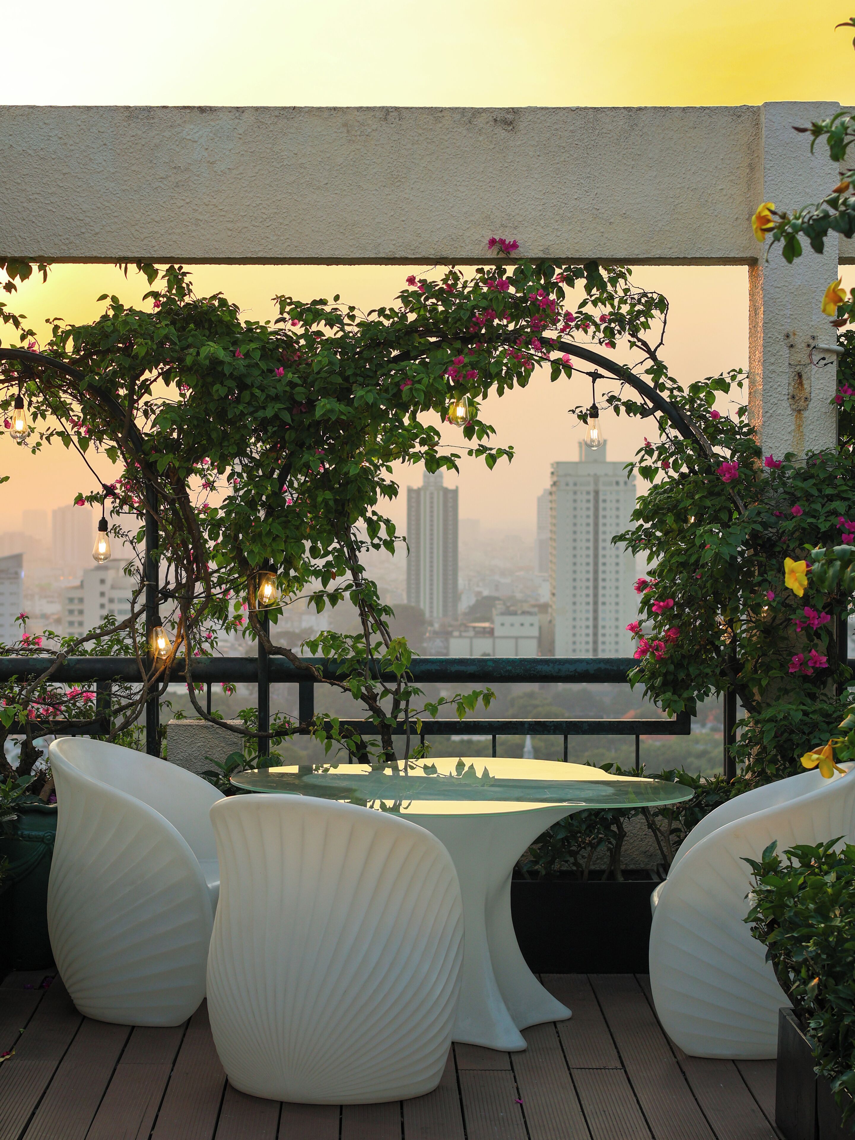 A terrace with a table and chairs, surrounded by greenery and flowers, with a cityscape in the background.