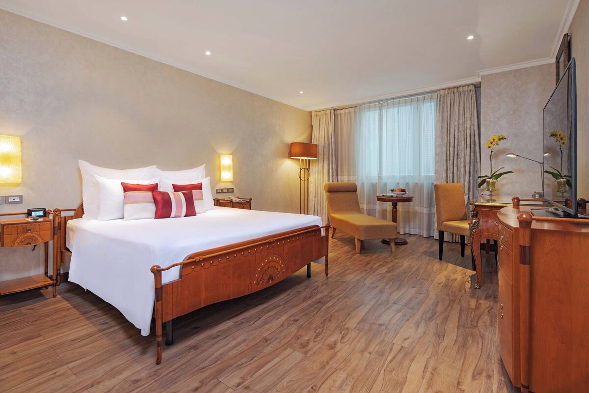 Une chambre d’hôtel avec un lit en bois, un bureau, une chaise, une lampe de bureau, un miroir et une fenêtre avec des rideaux.