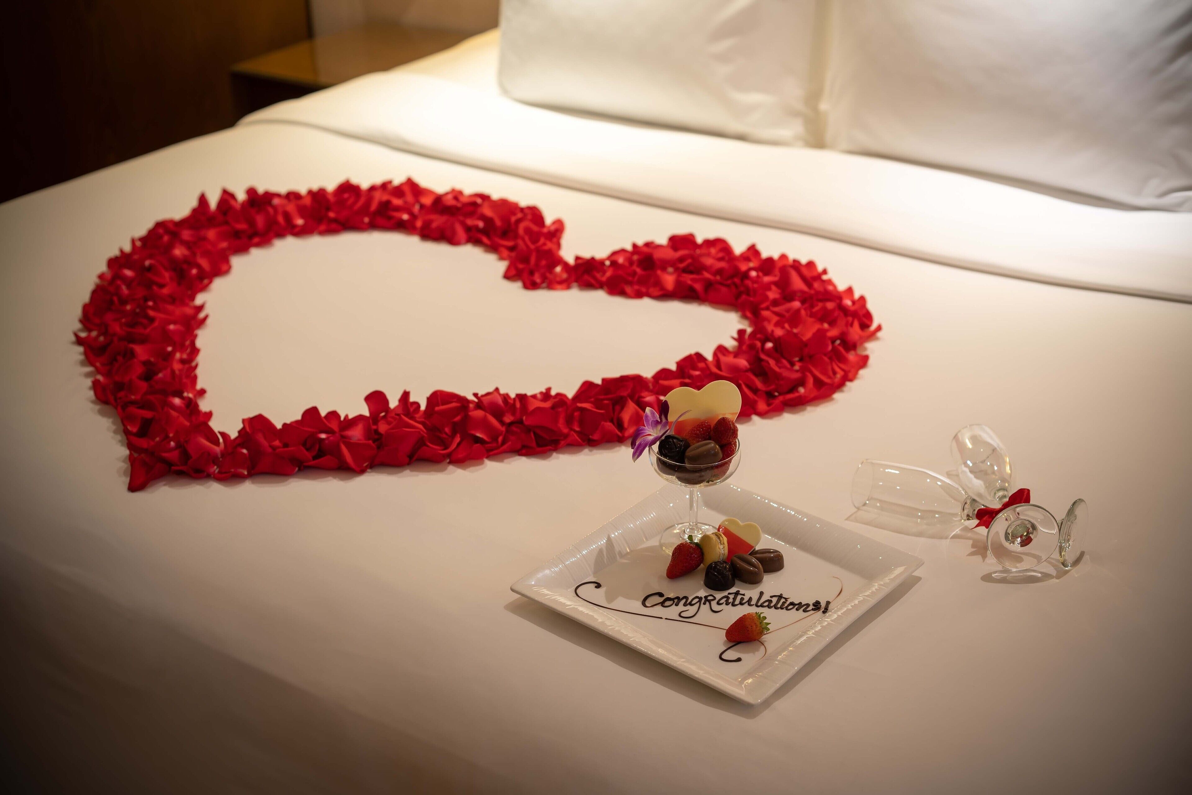 A bed with a heart-shaped arrangement of red rose petals and a small dessert plate with a heart-shaped decoration.