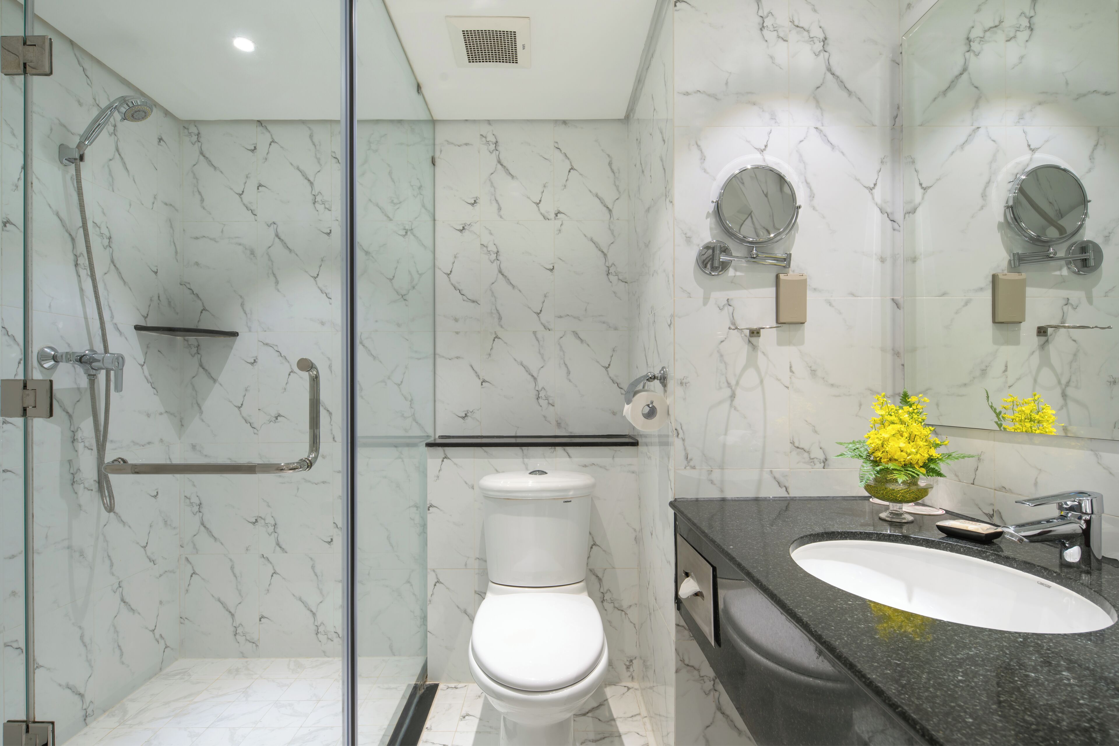 A modern bathroom with a glass-enclosed shower, a white toilet, a black countertop sink, and a mirror with a flower vase.