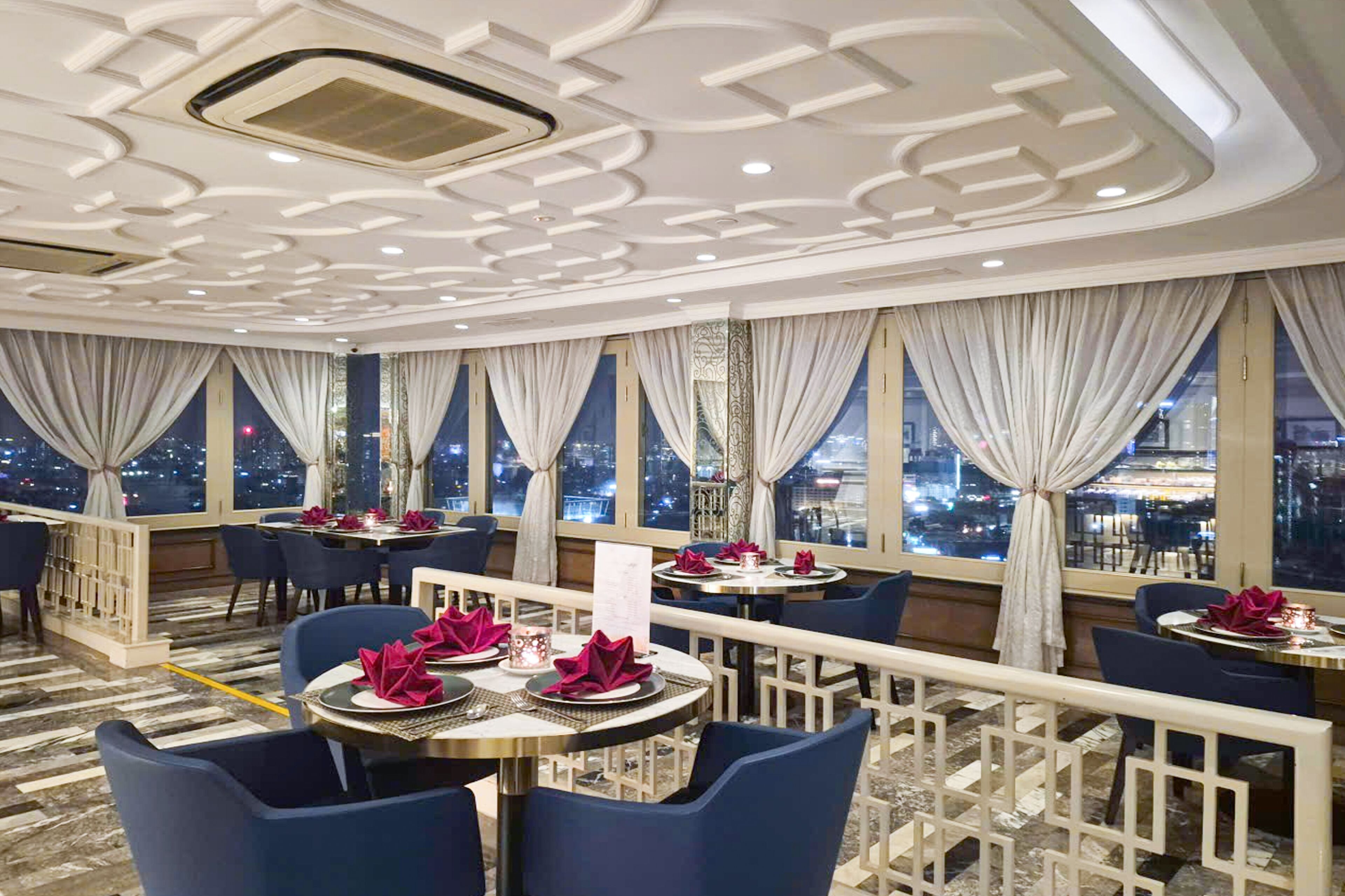 A restaurant with tables set for dining, large windows with curtains, and a city view at night.