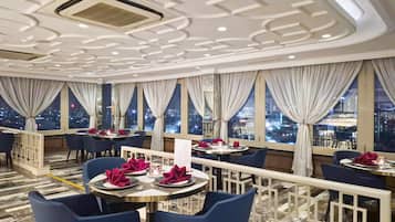 Een restaurant met gedekte tafels, grote ramen met gordijnen en uitzicht op de stad 's nachts.