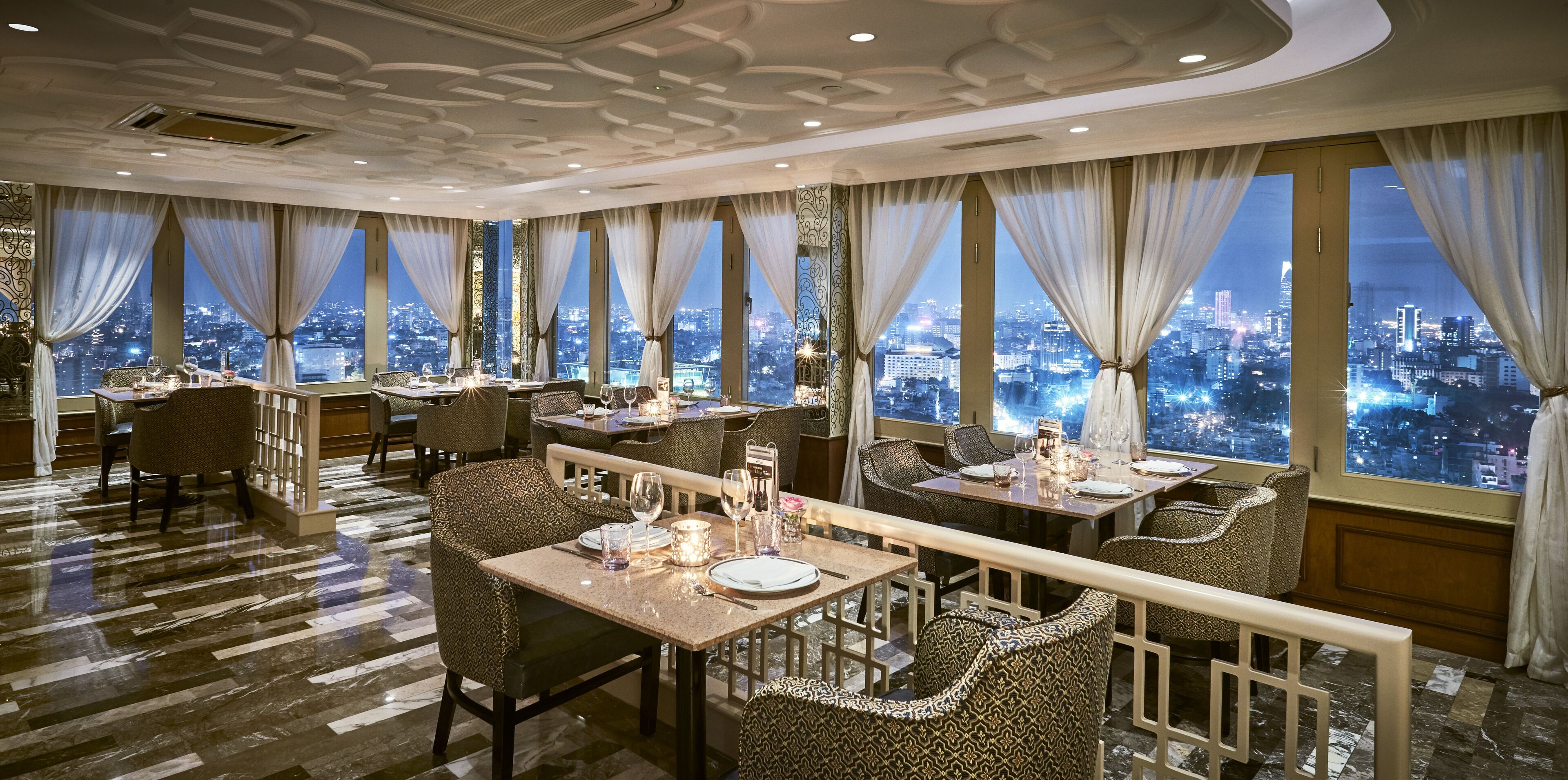 Een restaurant met gedekte tafels, grote ramen met gordijnen en uitzicht op de stad.