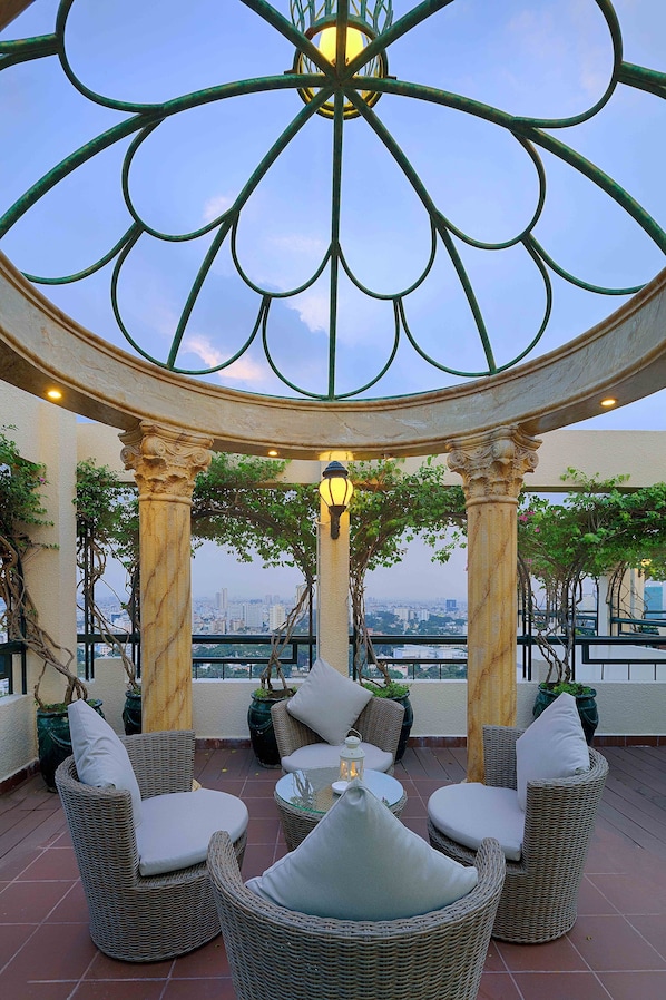 A rooftop terrace with wicker furniture, a decorative metal ceiling, and a city view.