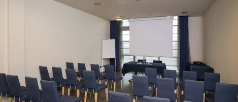 Un salón de conferencias con filas de sillas, un proyector, una pizarra y una mesa con un ordenador portátil.
