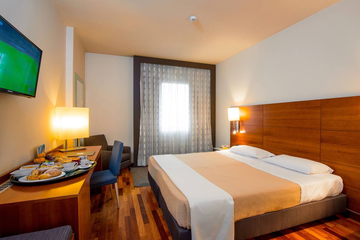 A hotel room with a bed, a desk with breakfast, a TV, and a window with curtains.