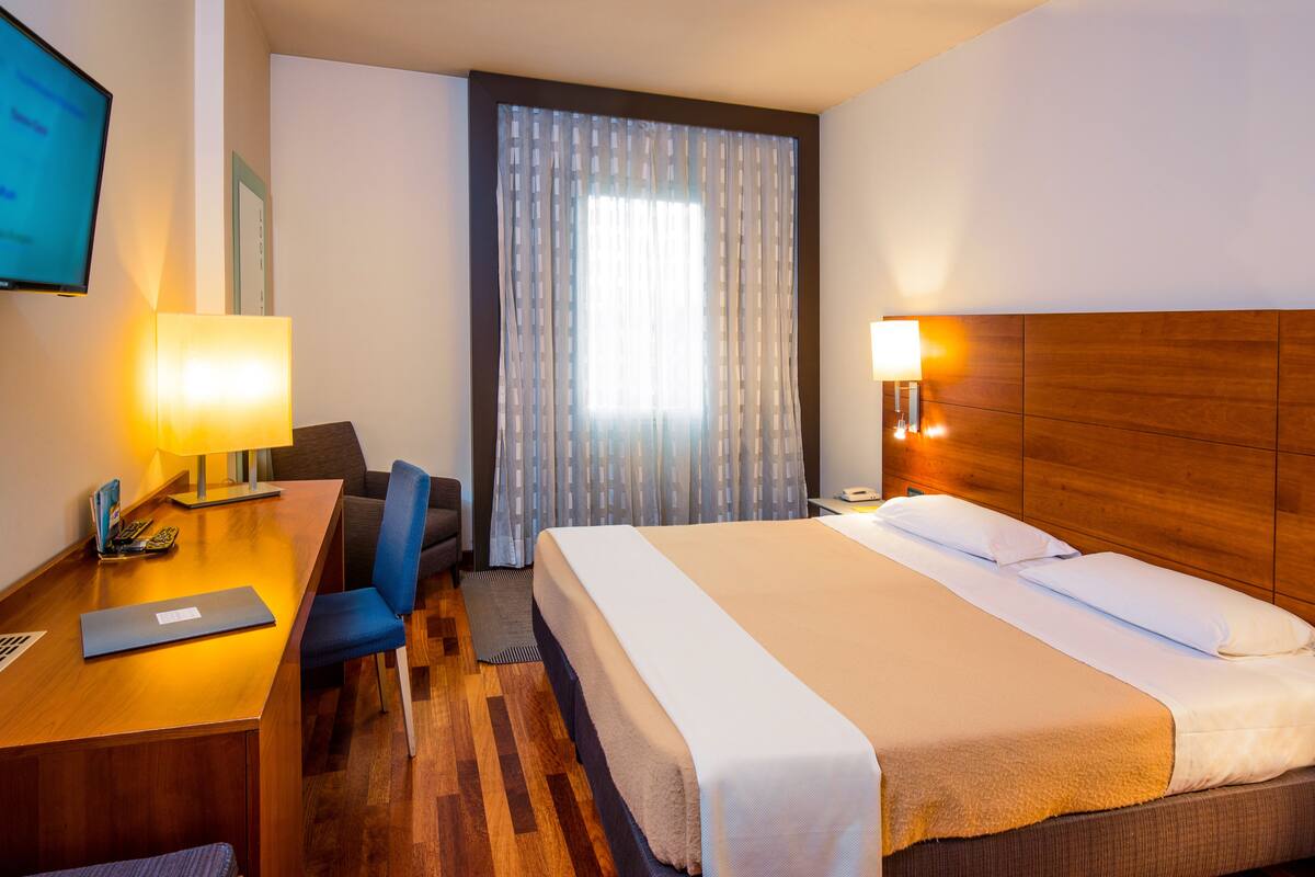 A hotel room with a bed, desk, chair, and a TV on the wall.