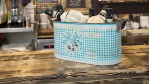 A bar area with a blue checkered basket containing bottles and glasses, a chalkboard menu, and a variety of liquor bottles on shelves.