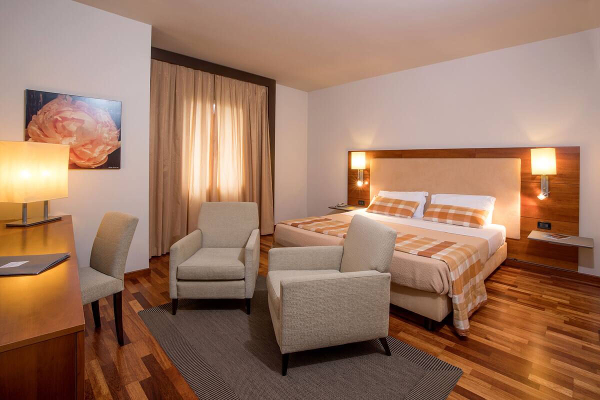 A hotel room with a bed, two armchairs, a desk, a lamp, and a painting on the wall.