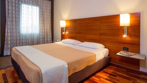 A hotel room with a large bed, wooden headboard, two bedside lamps, a nightstand, and a window with curtains.