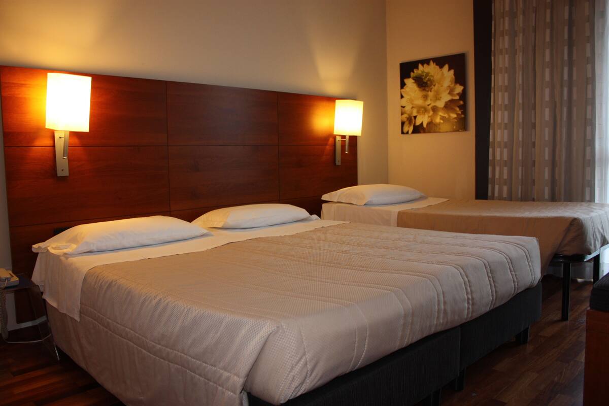 A hotel room with three beds, each with white bedding and headboards. There are wall-mounted lamps on either side of the beds.