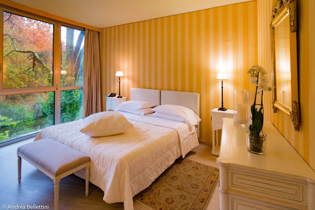 A bedroom with a large window, a bed with white linens, a bedside table with a lamp, and a mirror.