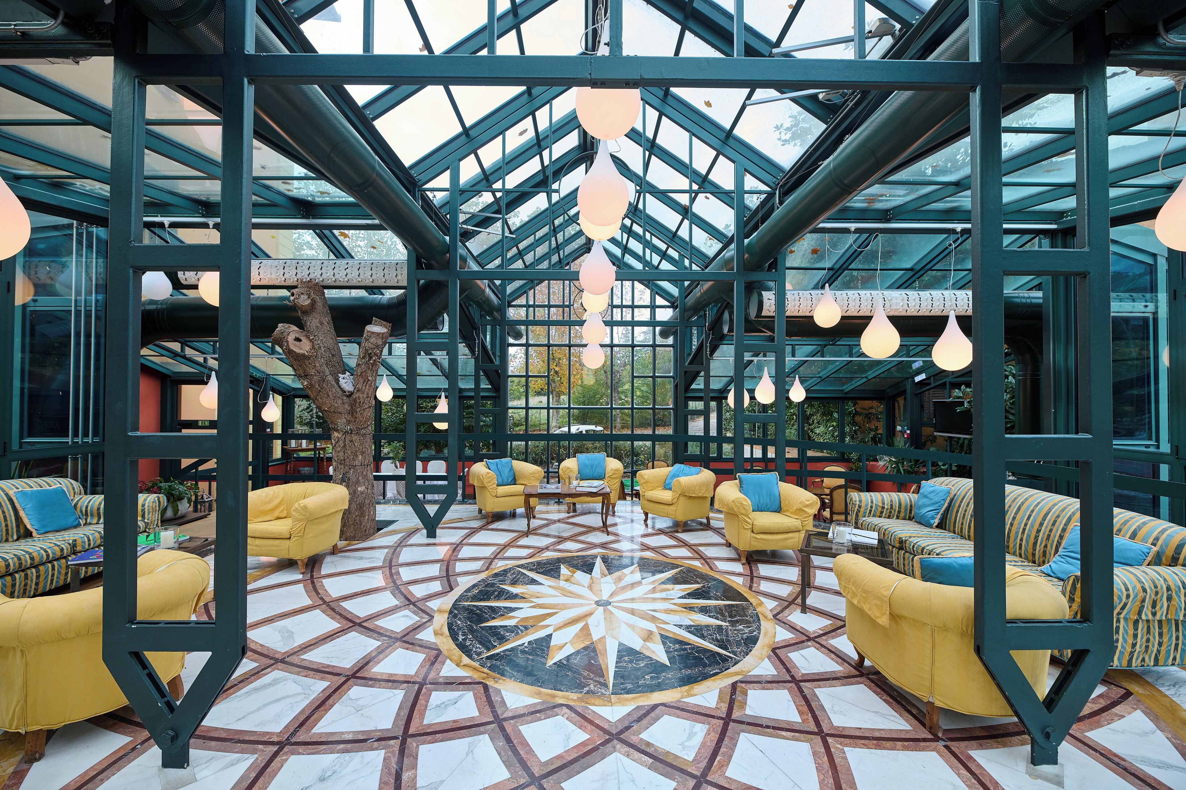 A spacious indoor area with yellow sofas arranged around a central geometric patterned floor, a large tree, and a glass ceiling structure.