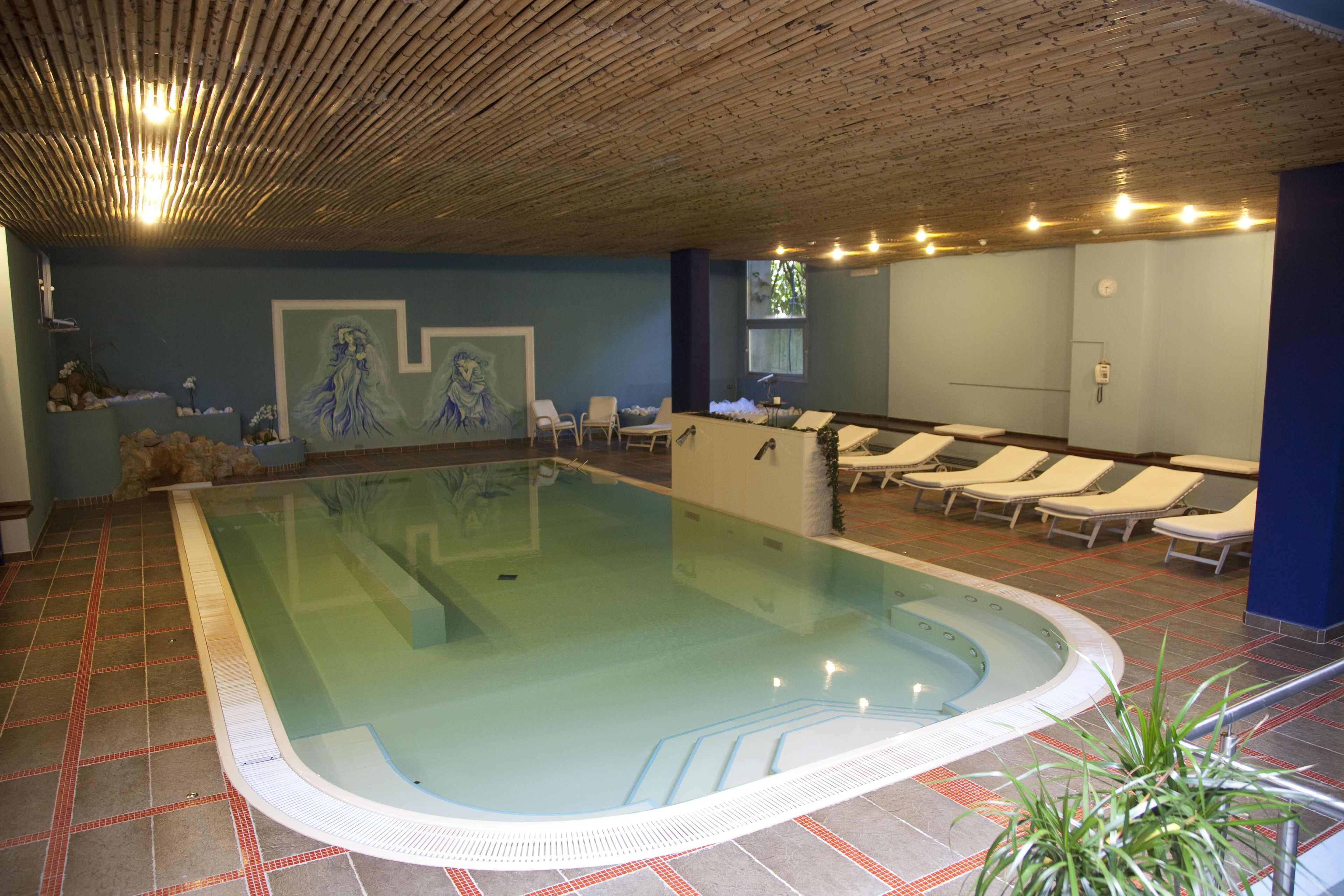 An indoor swimming pool with a tiled floor, lounge chairs, and a ceiling with wooden slats and lights.