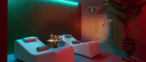 Une salle de spa moderne avec deux tables de massage blanches, une petite table avec une boisson et une plante en pot.