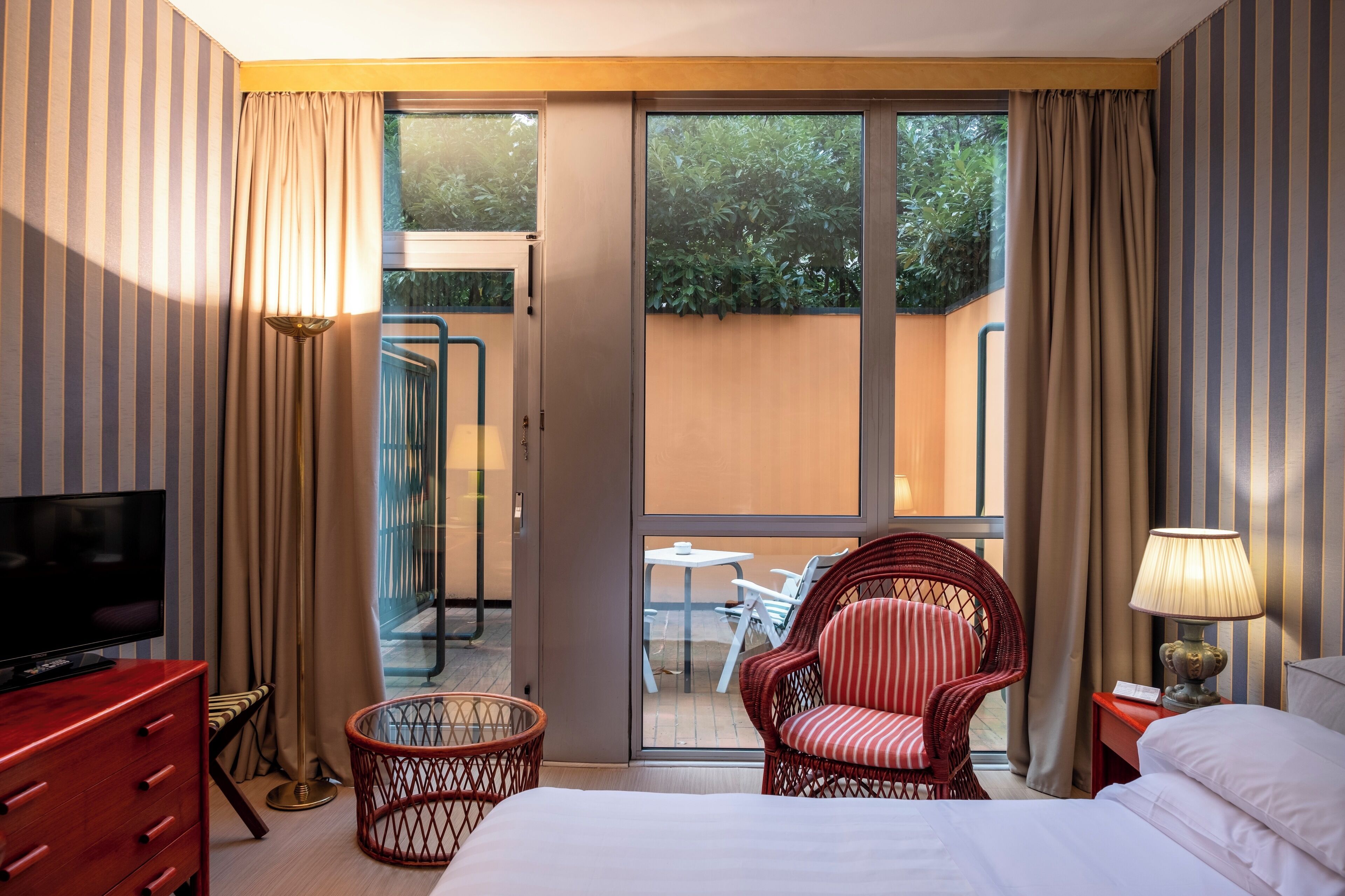 A hotel room with a bed, a television, a wicker chair, a small table, and a balcony with a view of greenery.