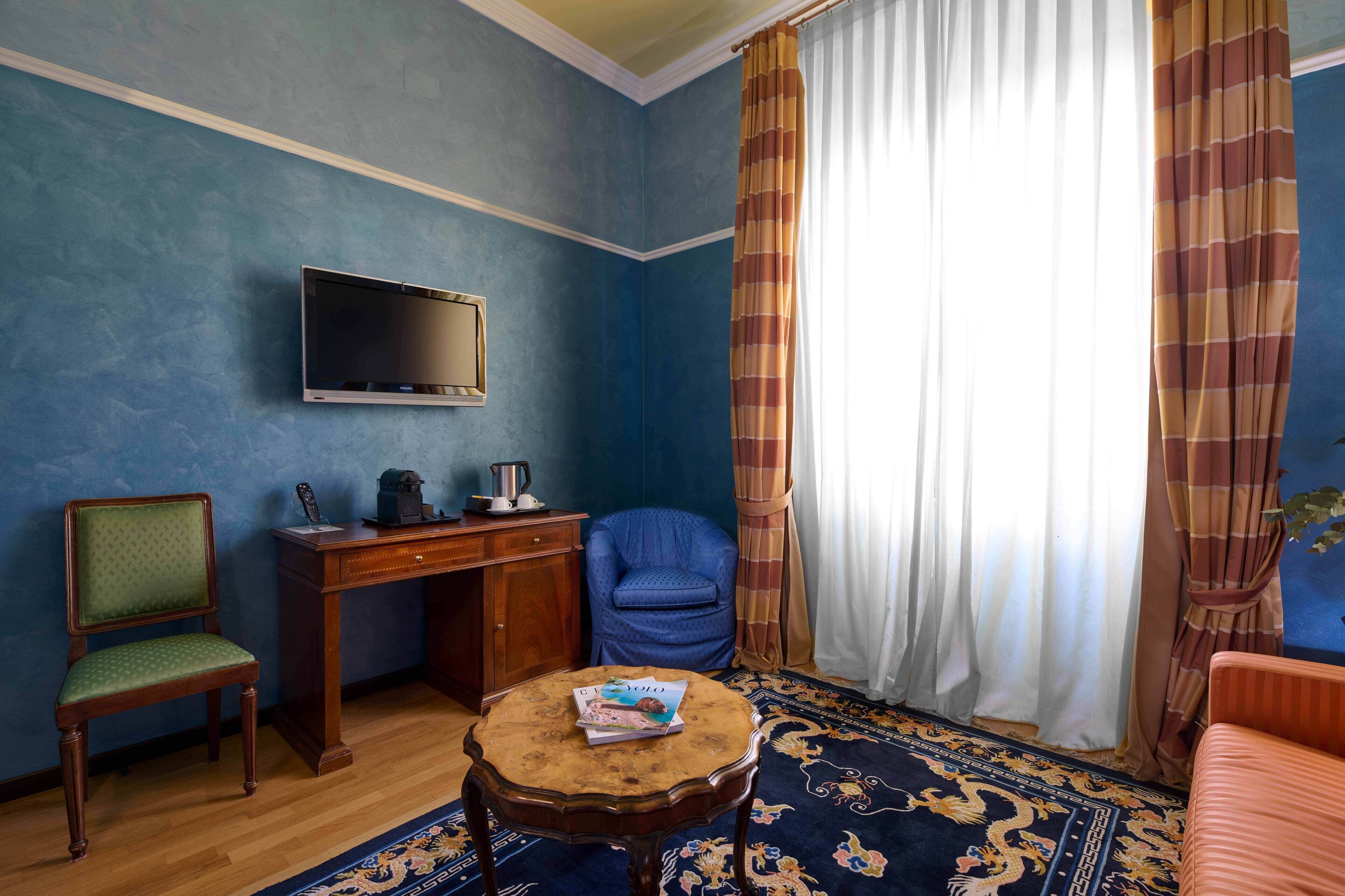 A room with a television mounted on the wall, a wooden desk with a chair, a round coffee table with a magazine, and a patterned rug.