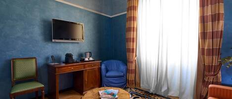 A room with a television mounted on the wall, a wooden desk with a chair, a round coffee table with a magazine, and a patterned rug.