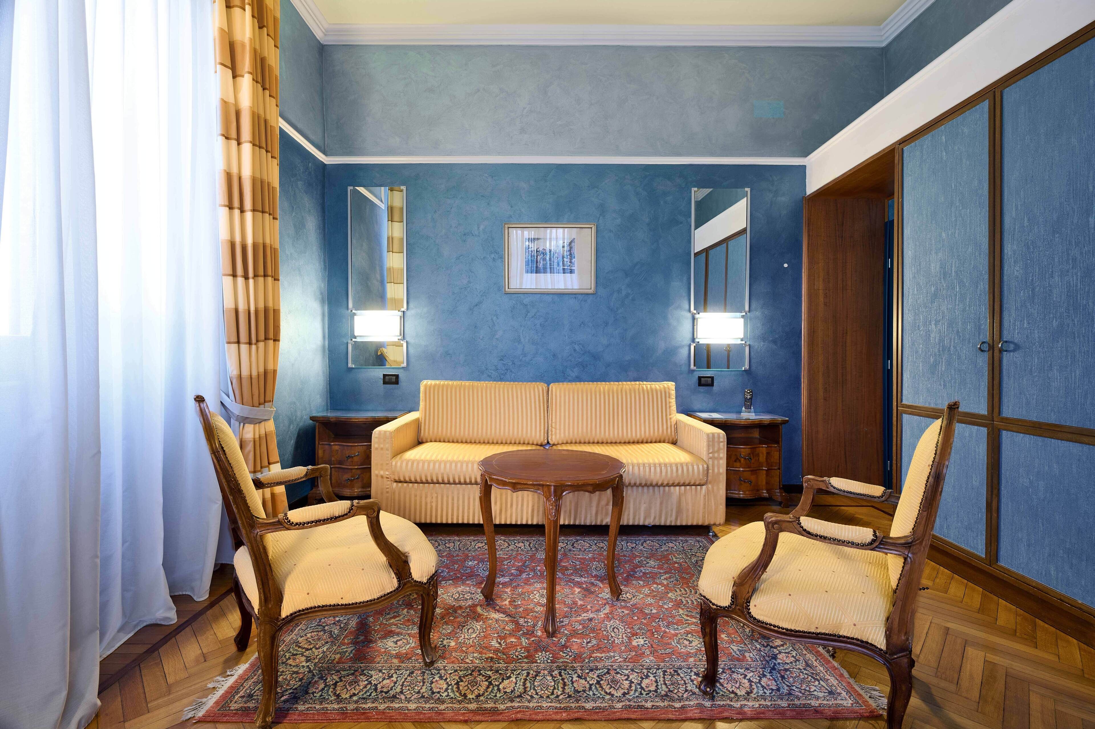 A room with a blue wall, a beige sofa, two wooden chairs, a small round table, and a patterned rug.