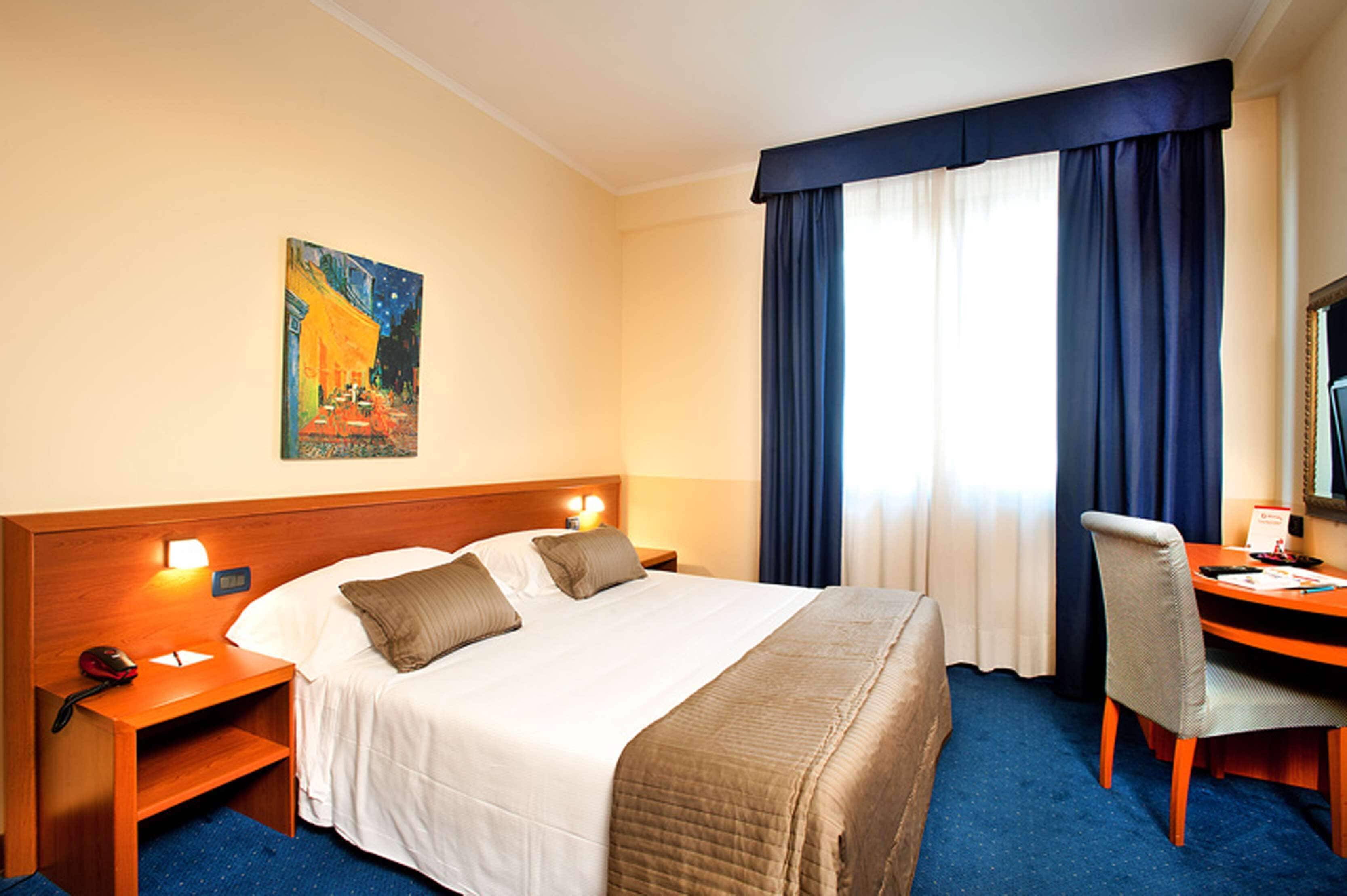 A hotel room with a large bed, two bedside tables, a desk with a chair, a painting on the wall, and a window with curtains.