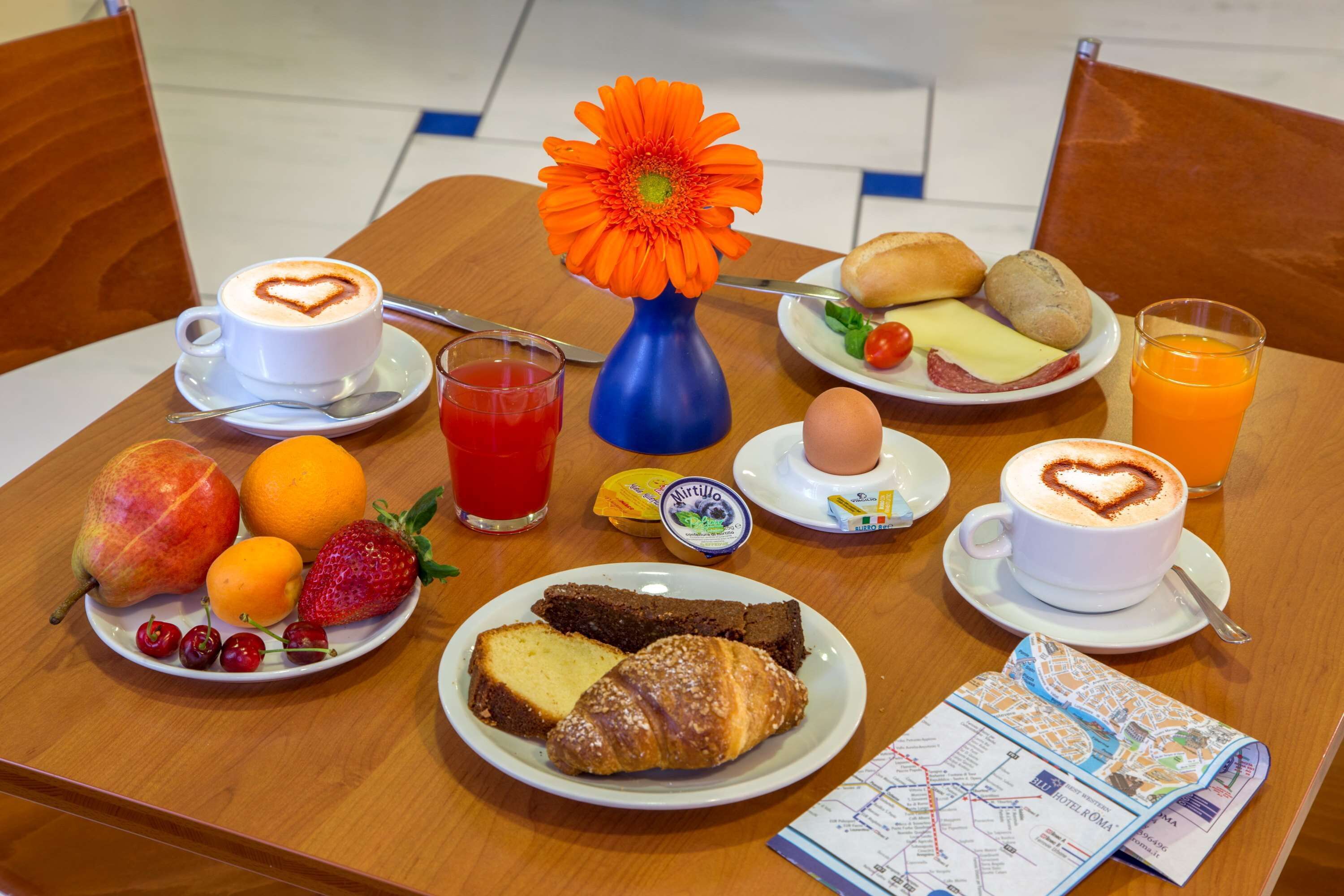 Una mesa de desayuno con una variedad de alimentos, entre ellos frutas, pasteles y bebidas, junto con un mapa y una flor decorativa.