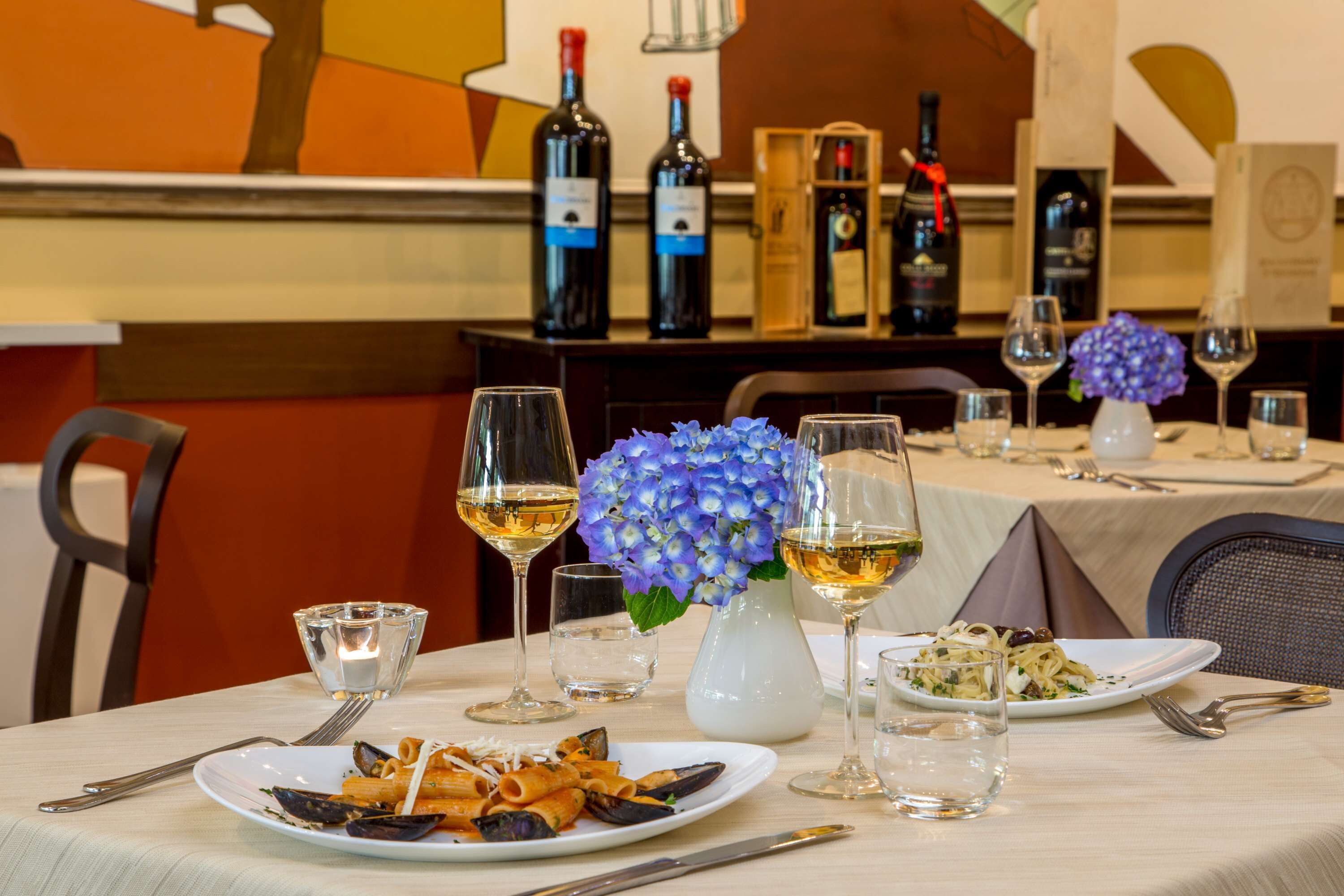Une table à manger avec des assiettes de pâtes et de fruits de mer, des verres à vin et un vase rempli de fleurs.