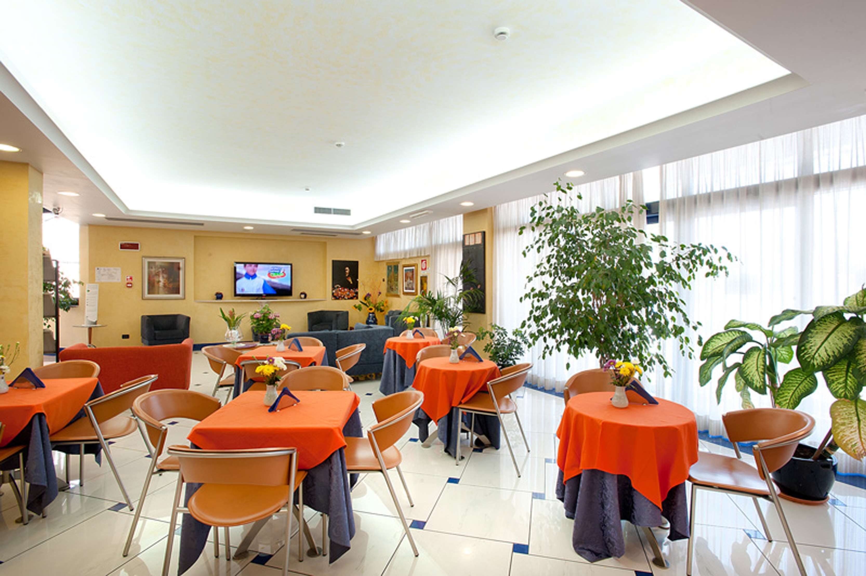 A hotel dining area with tables covered in orange tablecloths, chairs, and potted plants.