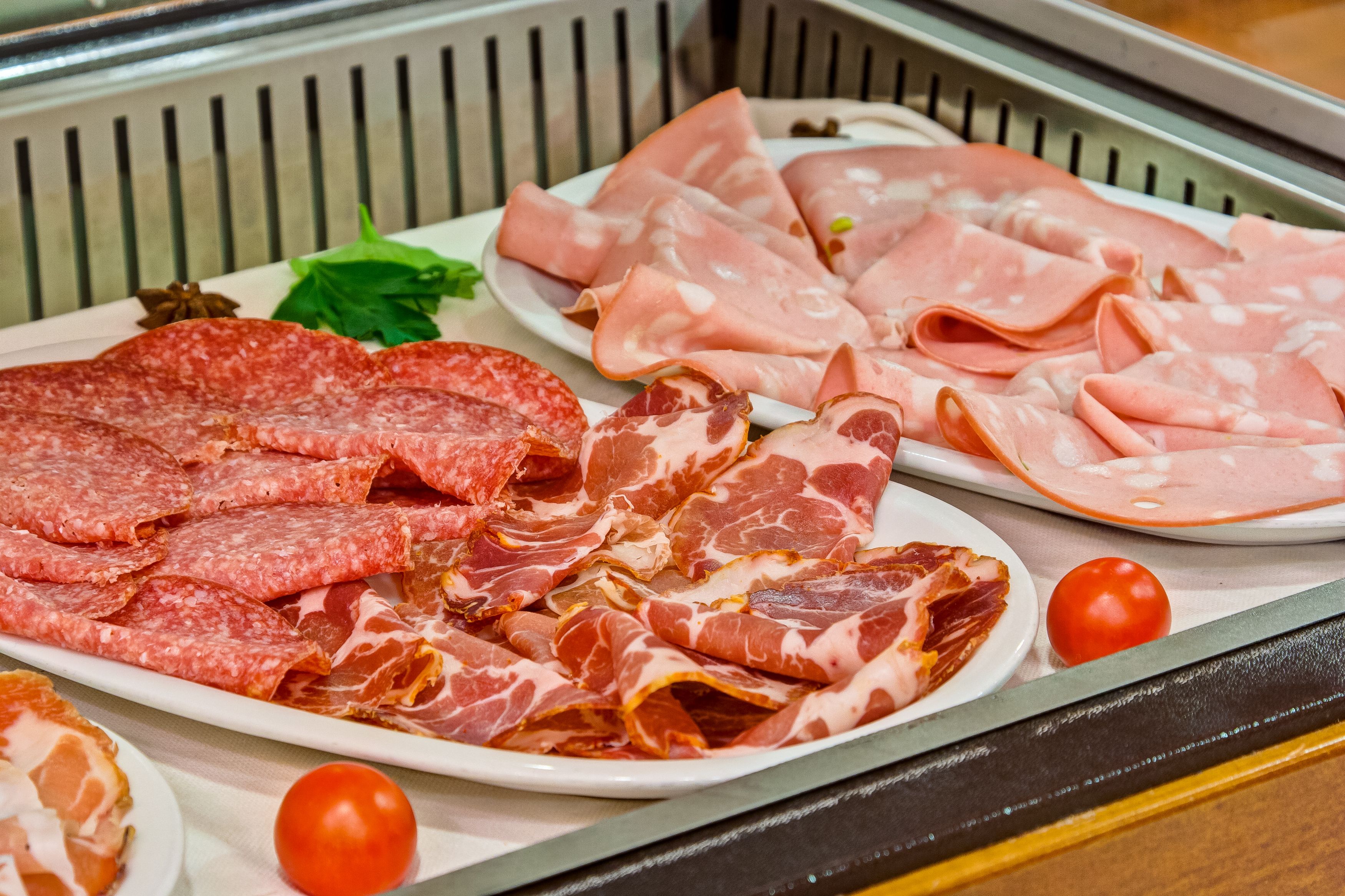 A variety of sliced meats on plates, including salami and prosciutto, with a tomato and some greens on the side.