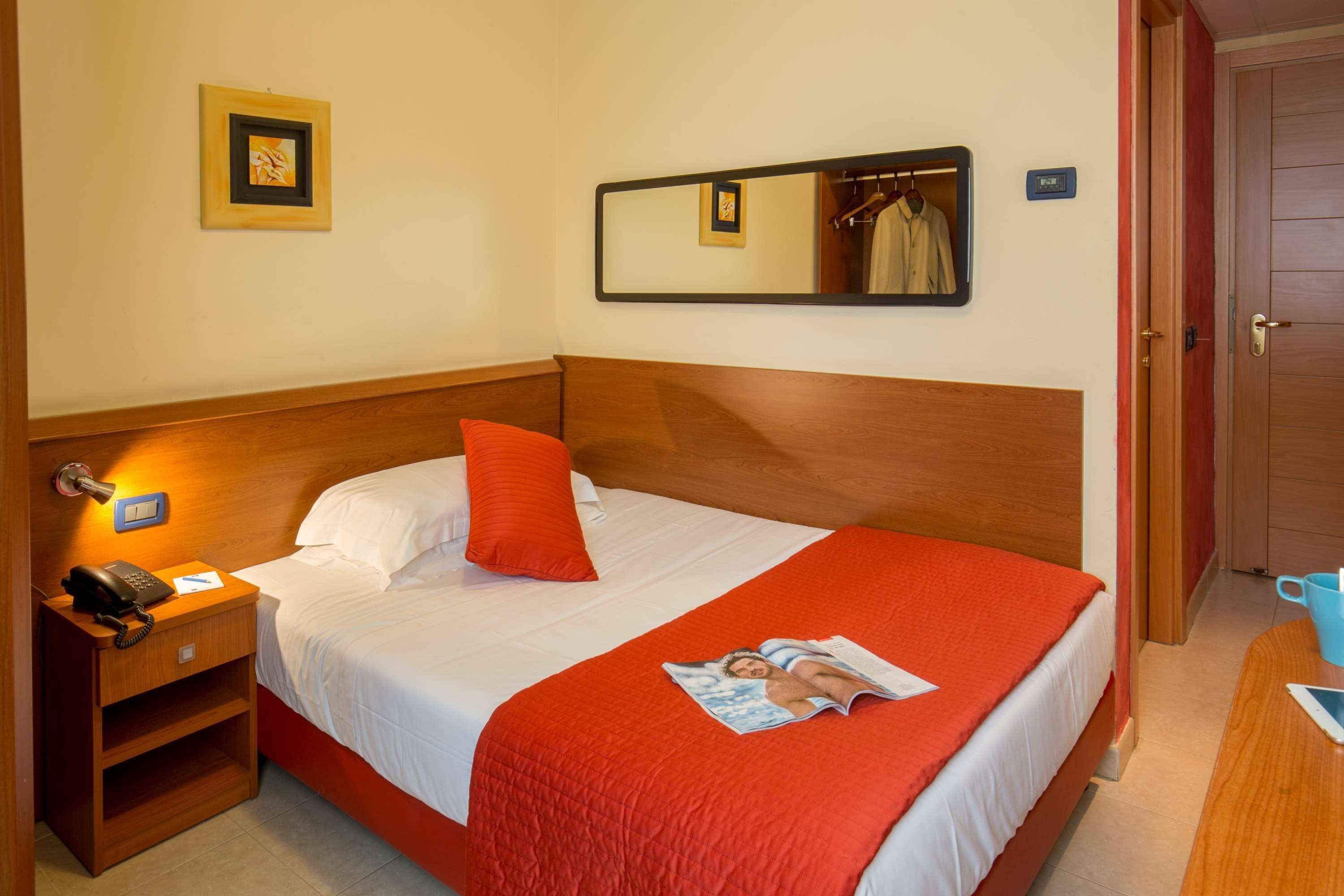 A hotel room with a wooden headboard, a single bed with white bedding and a red blanket, a bedside table with a phone and a lamp, a mirror on the wall, and a door to the right.