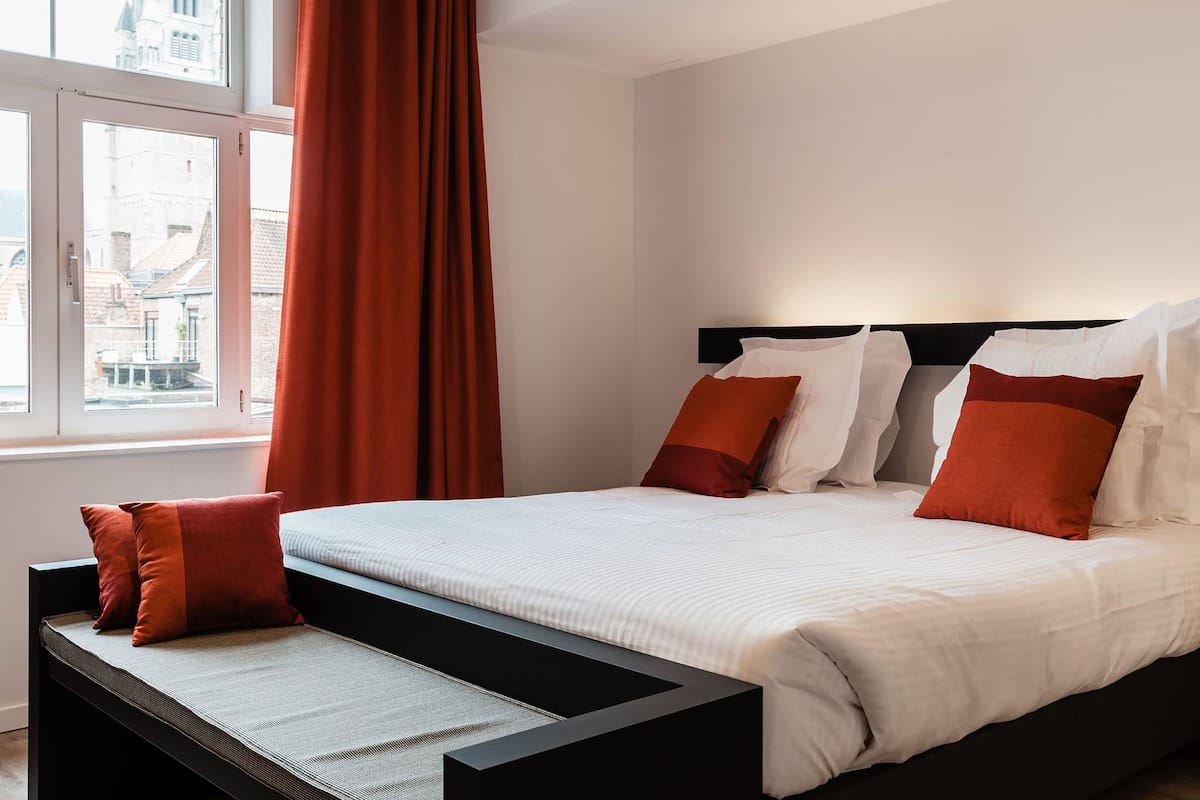 A hotel room with a bed, a window with curtains, and a view of buildings outside.