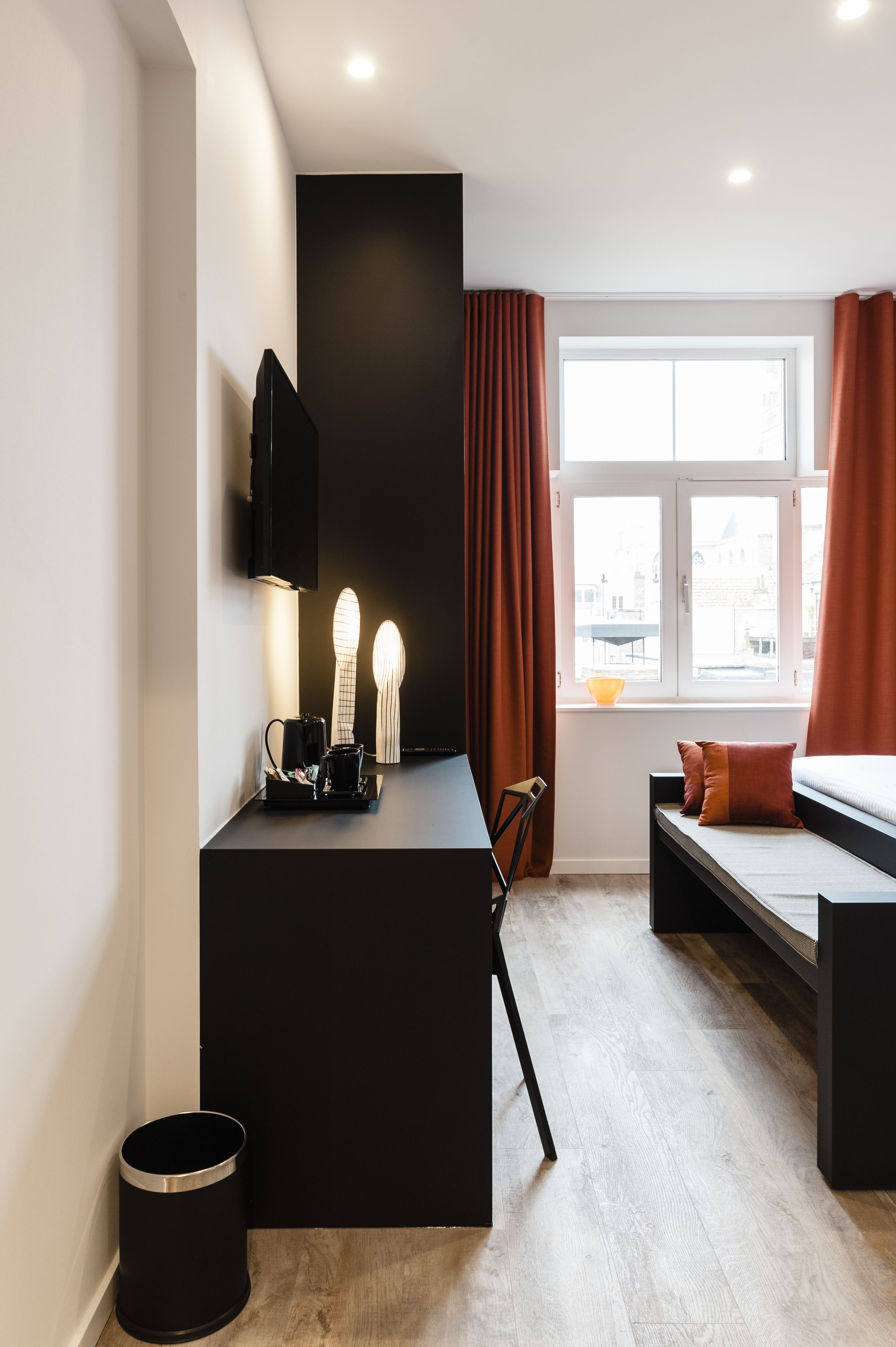 Una habitación moderna con un escritorio negro, una cama con almohada marrón, una ventana con cortinas y un televisor montado.